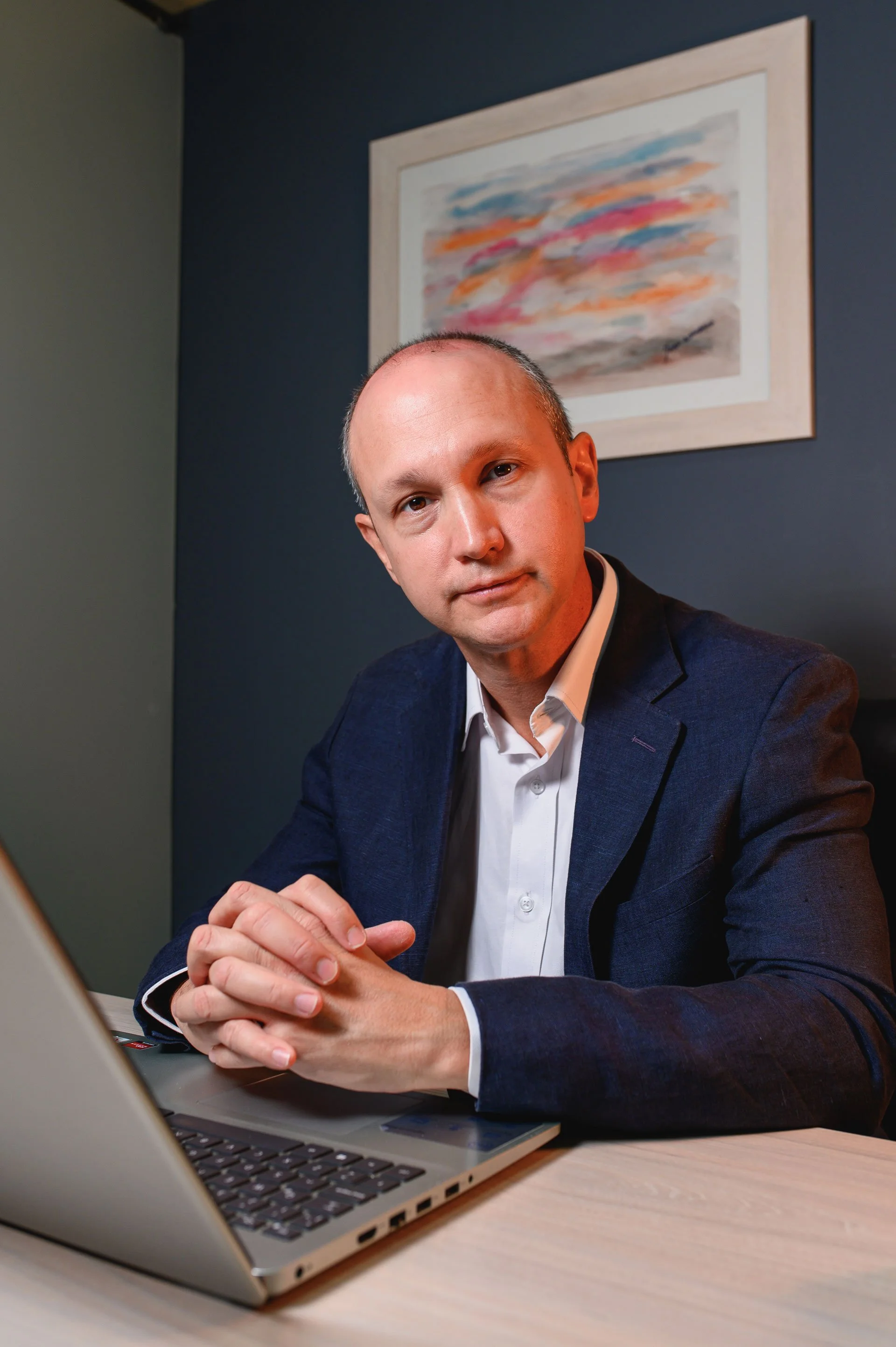 Hombre con ropa formal sentando frente a una computadora portátil, en una sala con paredes de colores oscuros y un cuadro con arte abstracto en la pared.