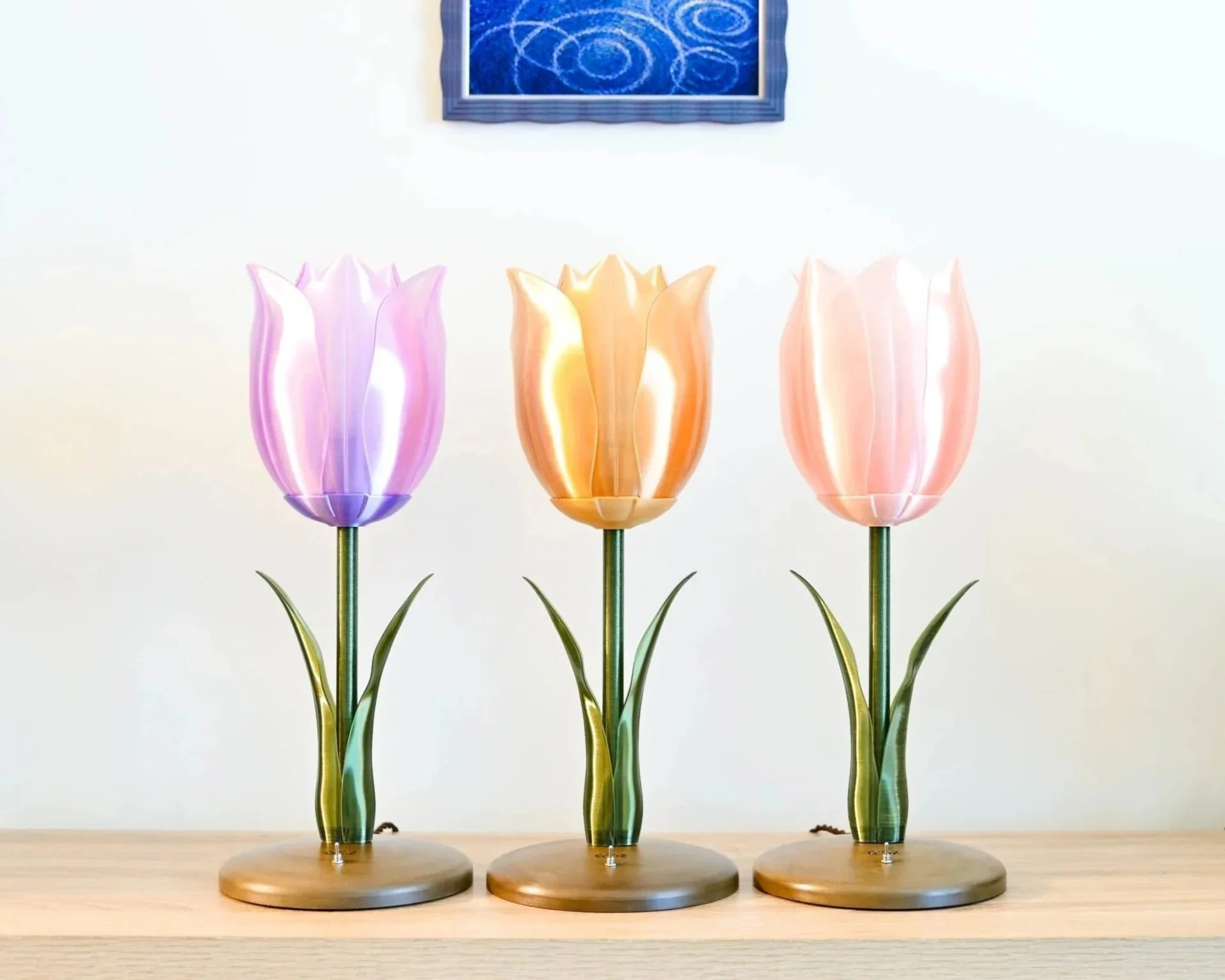 Three 3D printed table lamps in purple, orange, and pink, each on a round wooden base, with green stems and leaves, arranged in a row on a light wooden surface.
