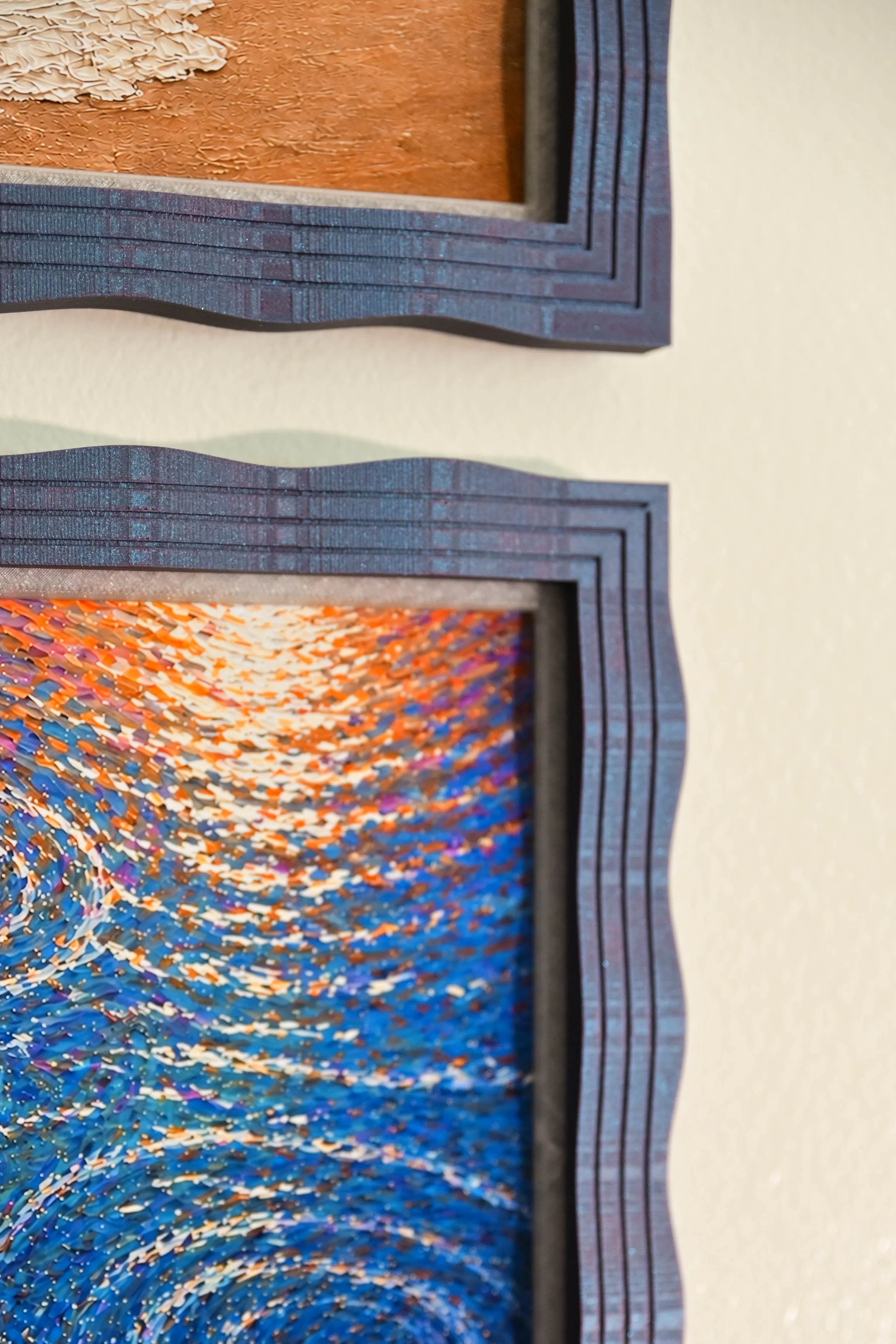 Close-up of a framed abstract artwork featuring a swirl of multicolored, textured pattern with shades of orange, purple, blue, and white. The frame is dark blue with layered, textured borders.