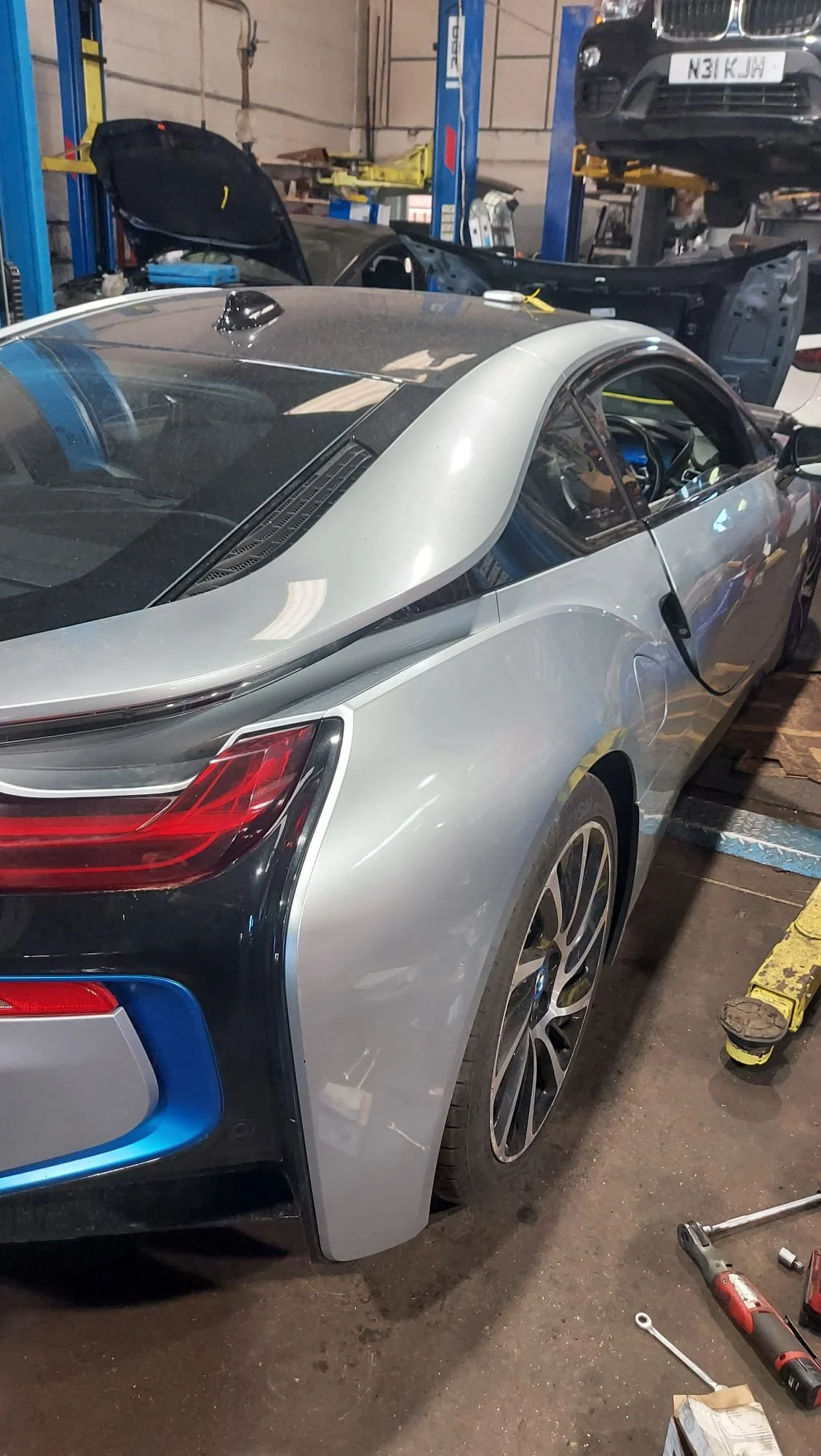 A silver BMW sports car in a garage or workshop undergoing repairs or maintenance. The car's rear and side are visible, with tools and equipment around it.