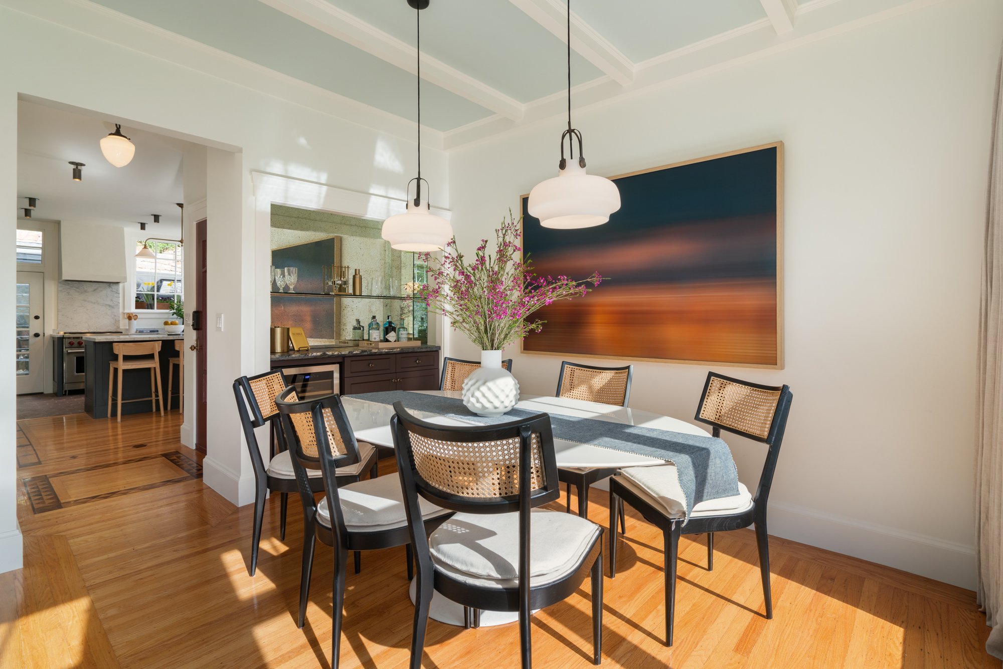 Dining room with a white round table, six chairs, a vase with pink flowers, a large abstract painting on the wall, and sunlight streaming in.