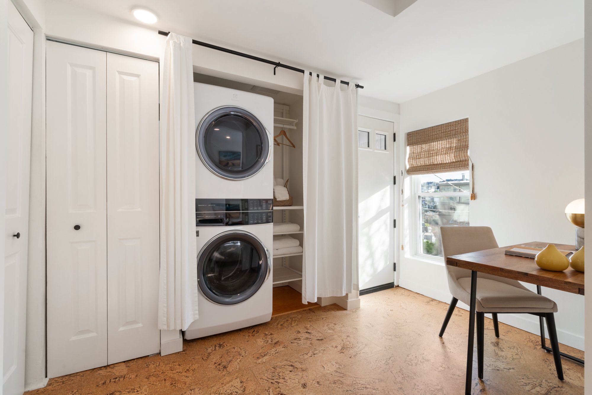 Laundry area with a stacked washer and dryer behind a white curtain, closet with shelves and folded towels, a window with a woven shade, and a small dining table with a chair and decorative vases