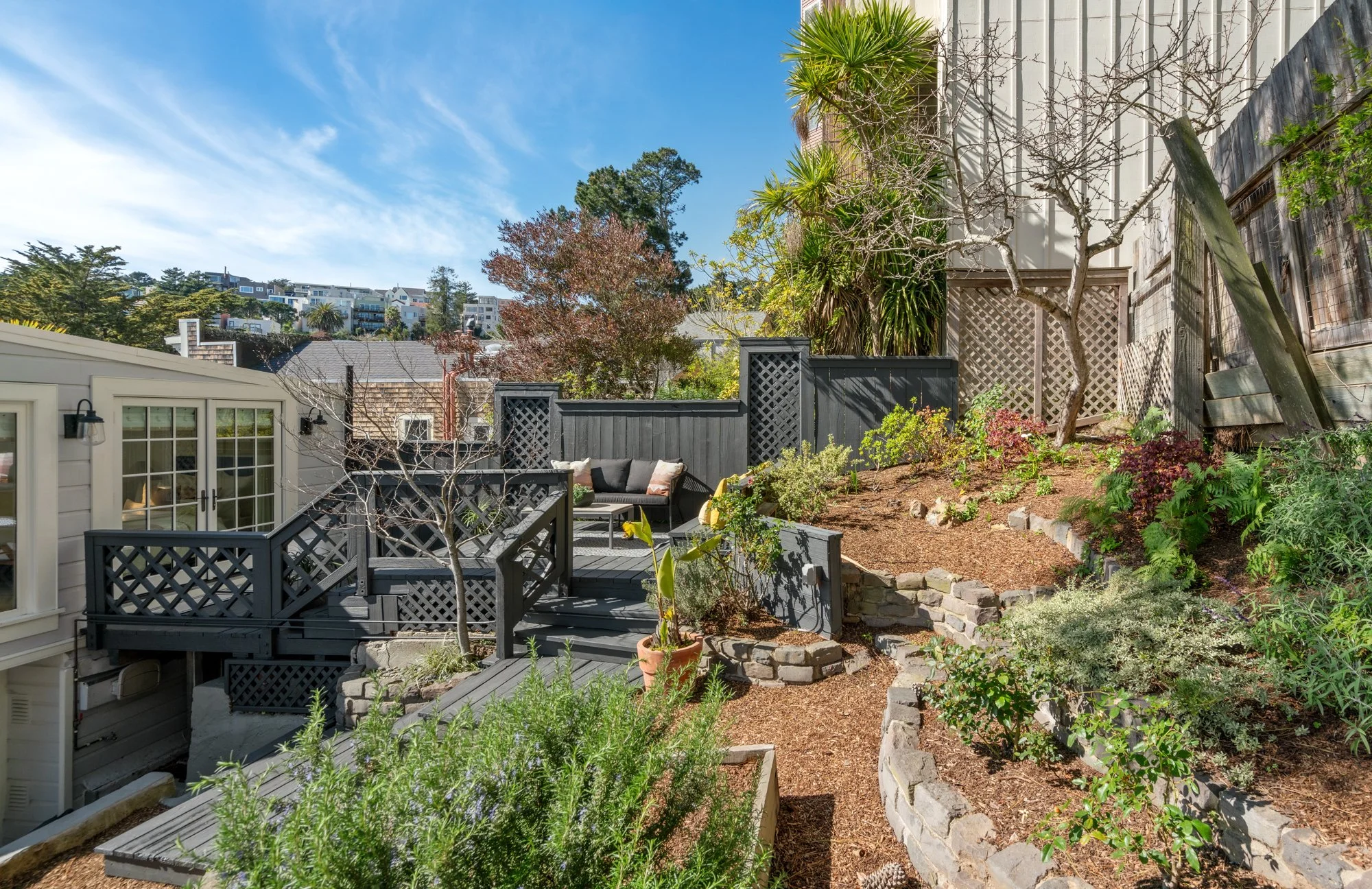 A backyard garden with a wooden deck, outdoor seating, and a variety of plants, trees, and shrubs, with a house, fencing, and buildings on a hillside in the background and a blue sky with clouds overhead.