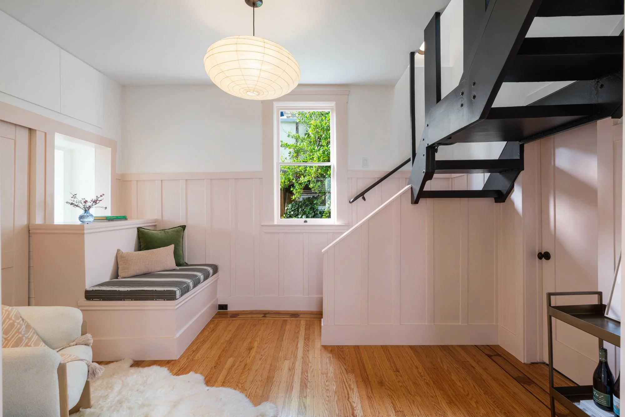 A cozy living space with wooden flooring, a window with lush greenery outside, a built-in bench with cushions, a black staircase, and a side table with a bottle and a frame.
