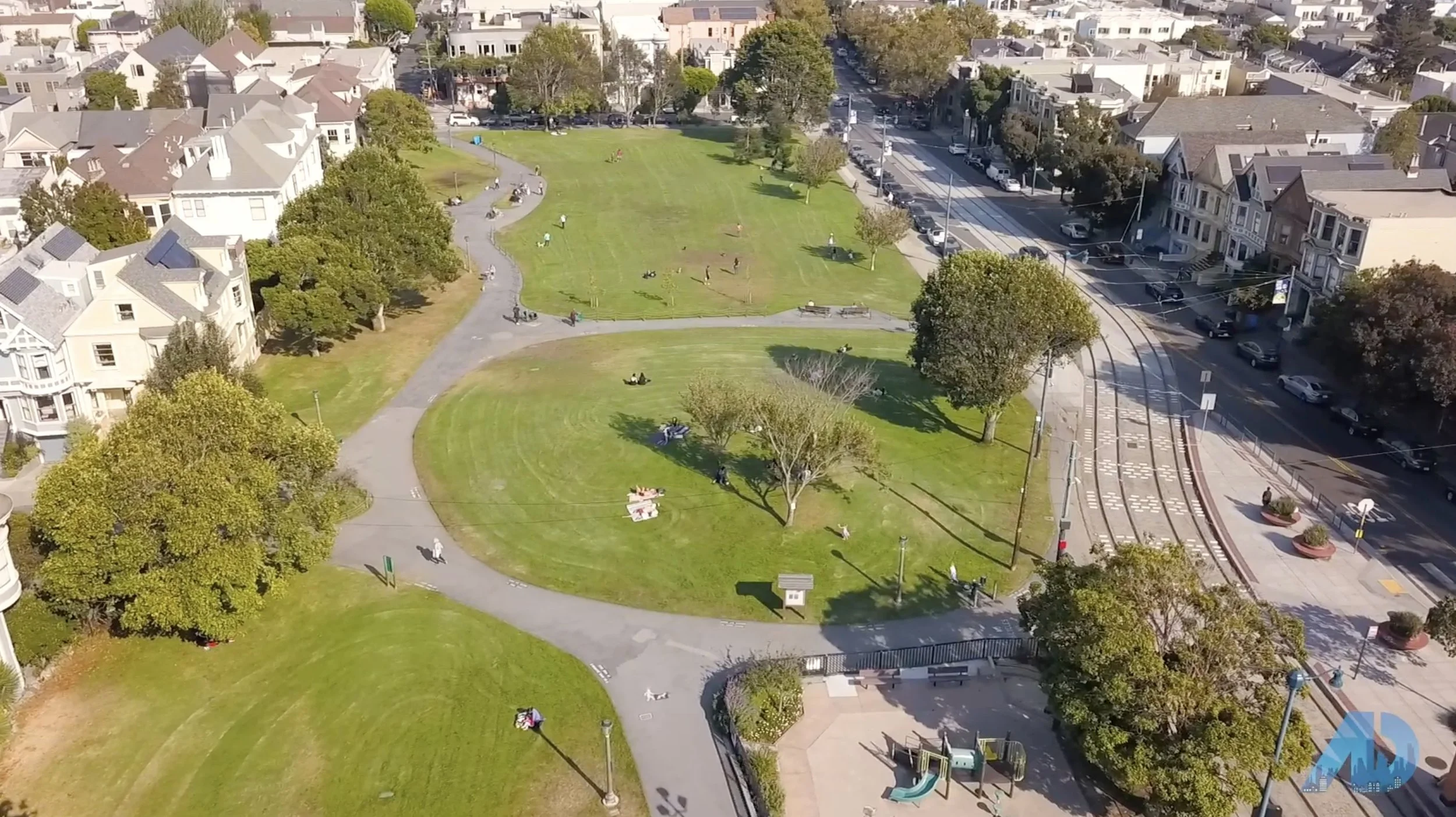 Aerial view of a city park with grassy areas, trees, walking paths, and people relaxing or playing; adjacent to a street with cars, tram tracks, and urban buildings.