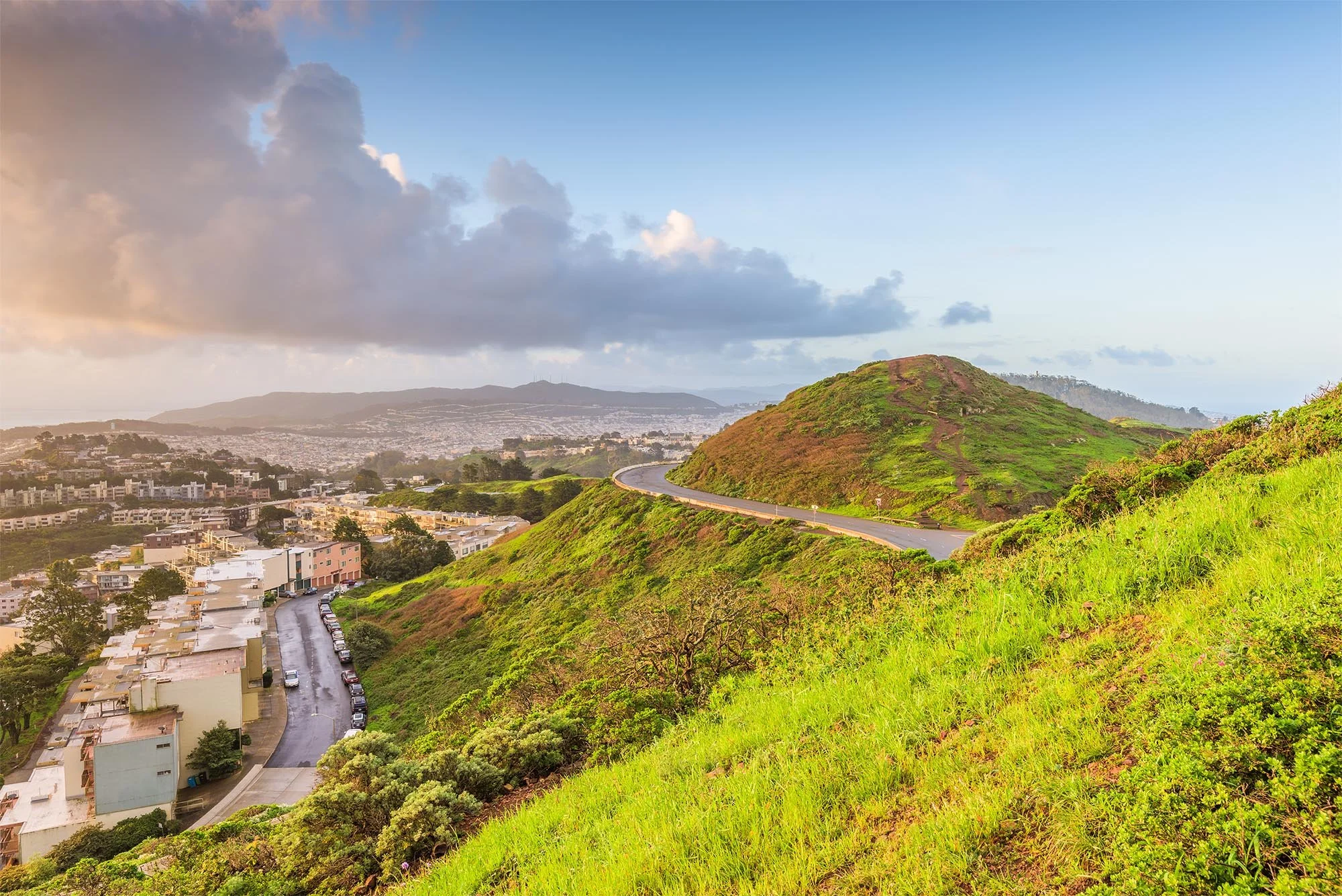 Scenic view of a green hillside with a winding road, overlooking a city with buildings and a distant mountain range, under a partly cloudy sky during sunset.