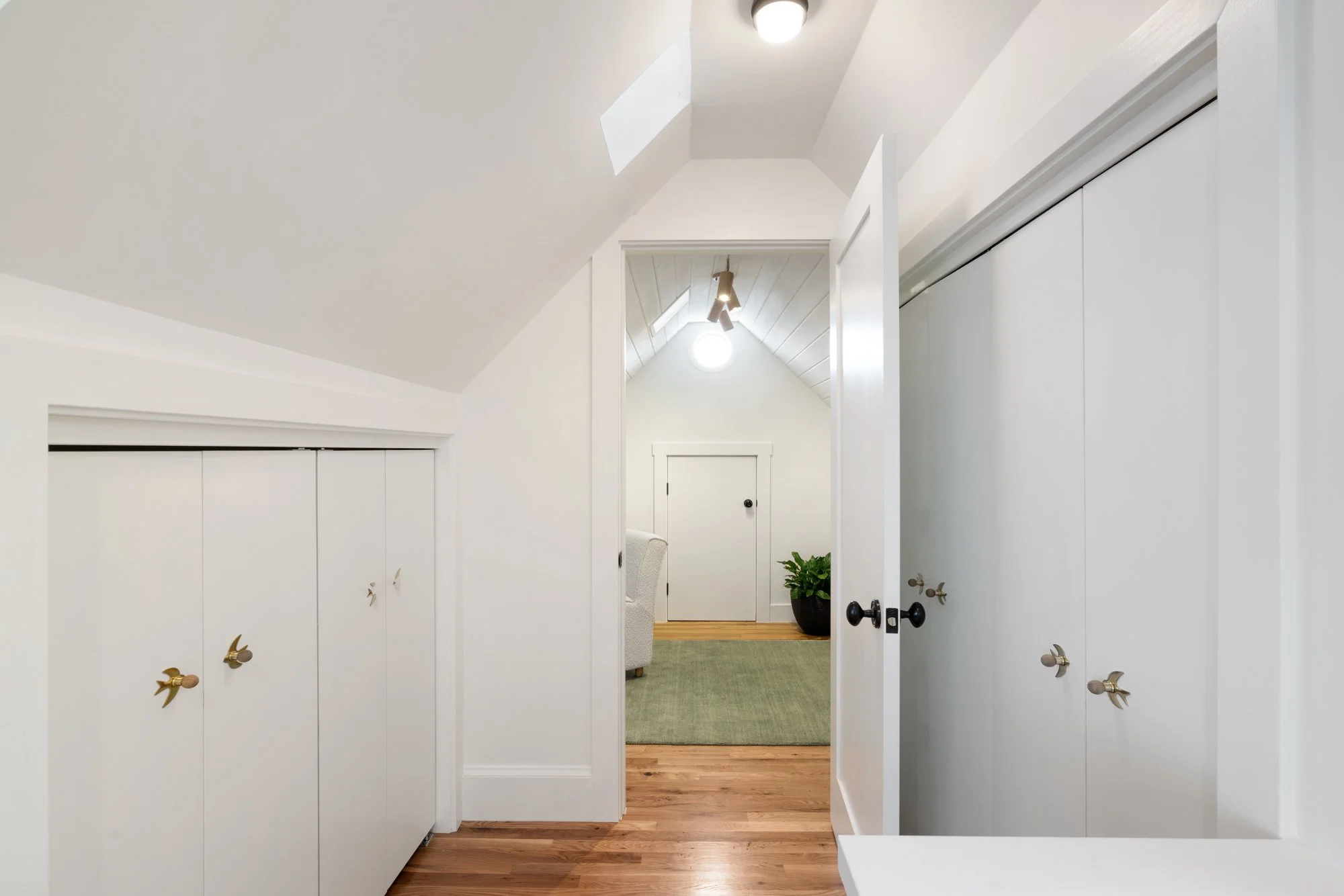 Interior view of a bright, white-walled attic room with built-in cabinets, hardwood flooring, and a door leading to a small space with a ceiling light, a potted plant, and a green rug.