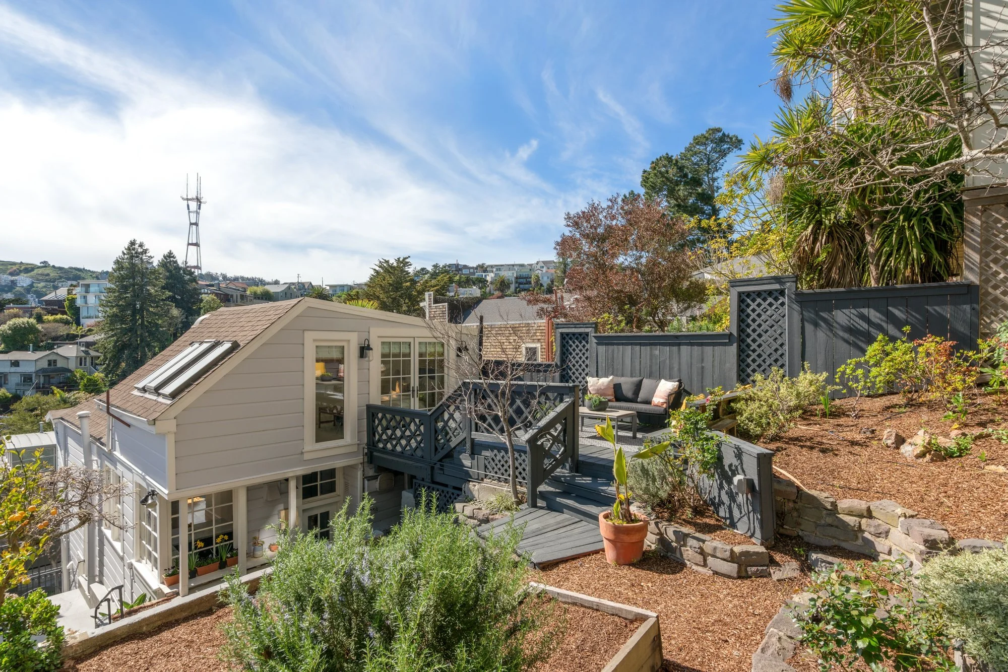 A backyard deck and garden with outdoor seating, plants, and a hillside view with houses and trees under a partly cloudy sky.