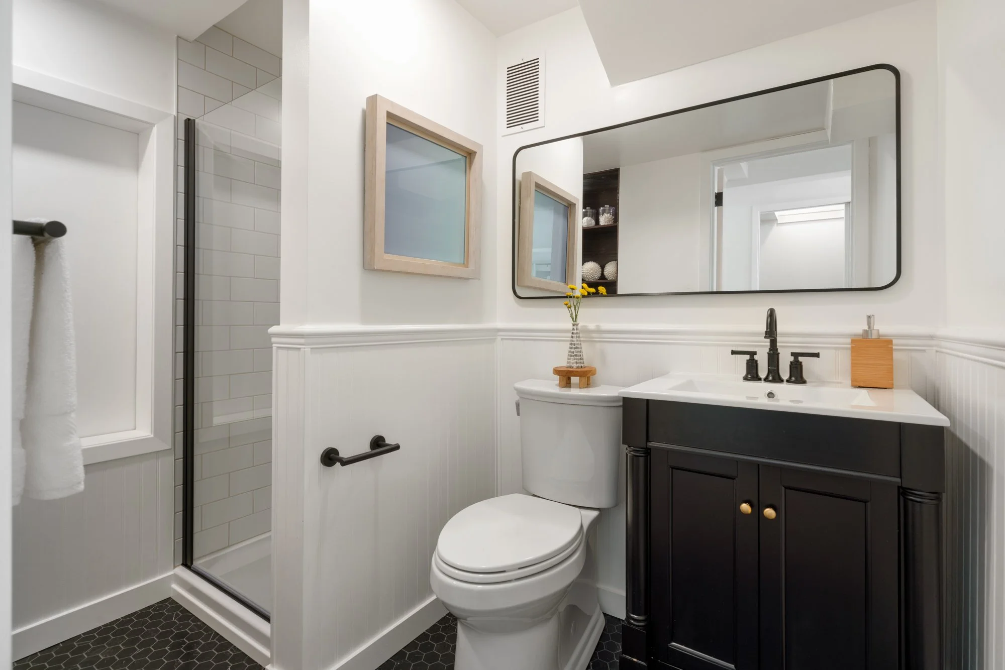 Modern bathroom with white walls, a black vanity with gold knobs, a white toilet with a small vase of yellow flowers on it, a large rectangular mirror, a small framed window, and a walk-in shower with white tile and black hardware.