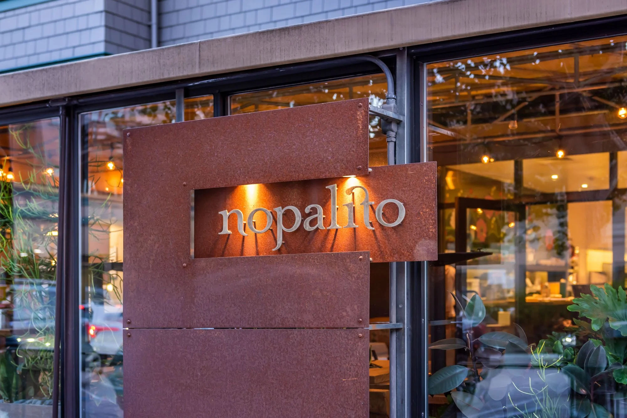 Restaurant sign reading 'Nopalito' with a rust-colored metal background, illuminated by warm lighting at the entrance, with glass windows showing a view of the interior.