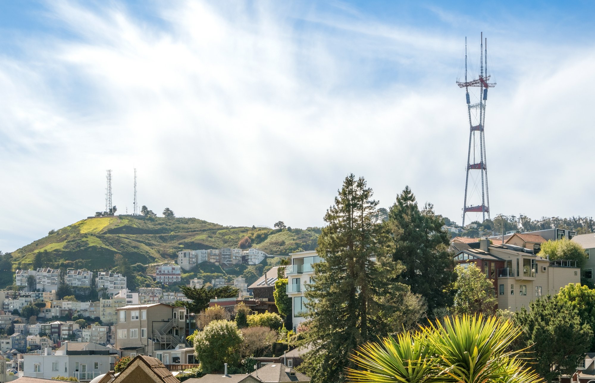 A scenic view of a residential area with houses and trees, with a hill in the background featuring communication towers and a large transmitter, under a partly cloudy sky.