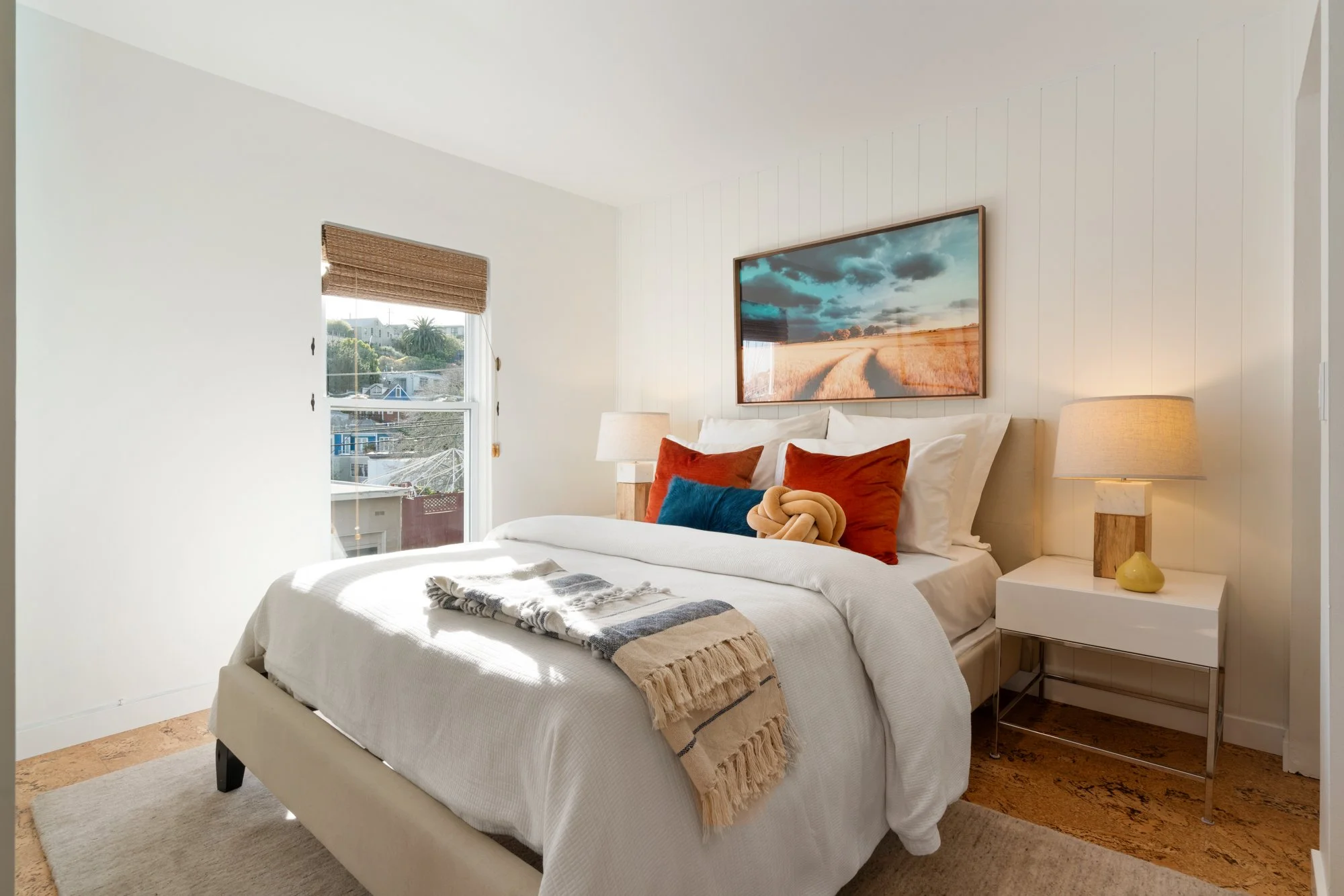 Bright bedroom with white walls, a large bed with white bedding and colorful pillows, two bedside lamps, a framed landscape photo above the bed, a window with a bamboo shade showing trees and houses outside.