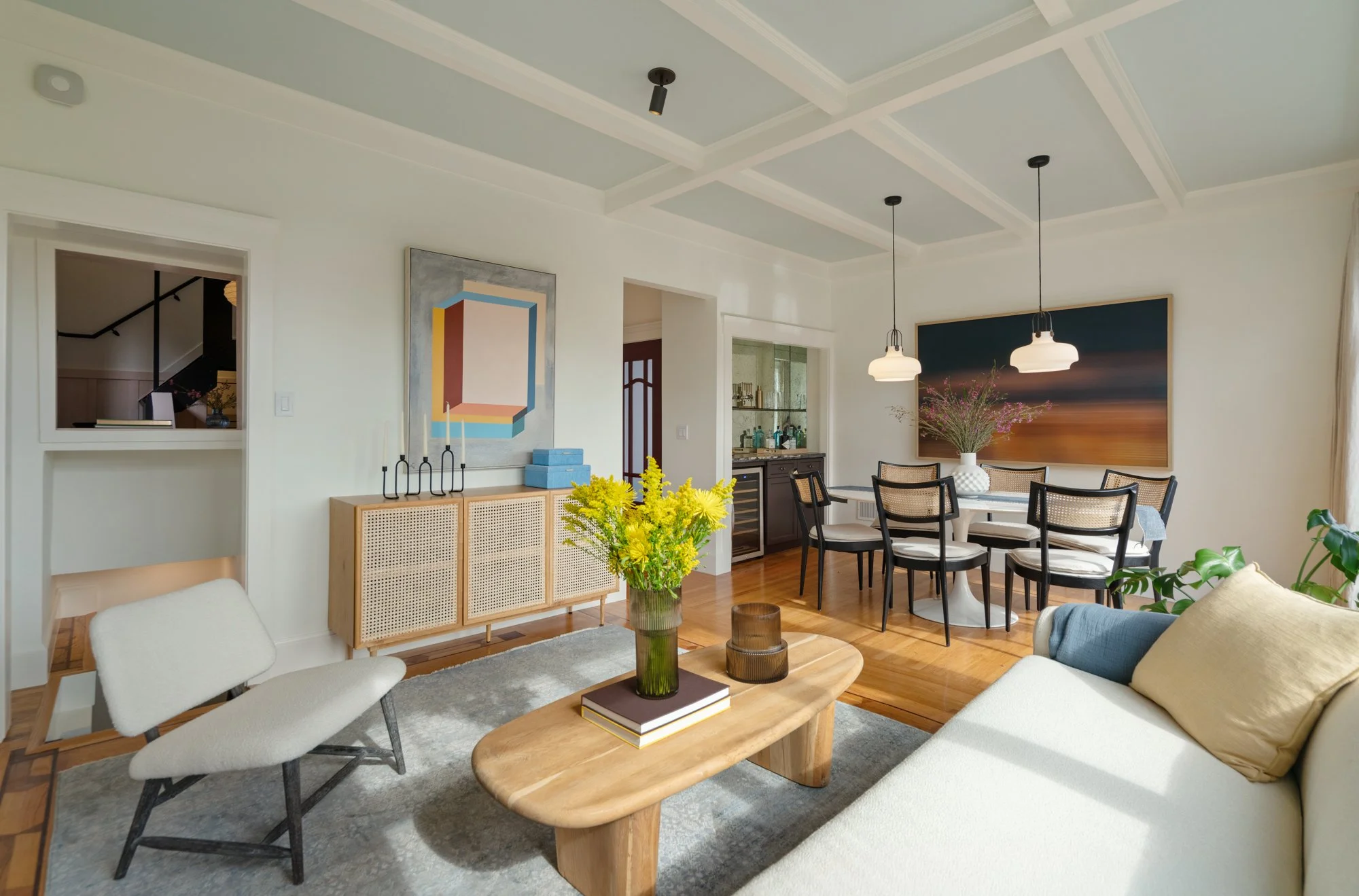 Living room with white walls, wooden floor, a beige sofa with cushions, a wooden coffee table with a vase of yellow flowers, two white chairs, a dining table with six chairs, and various artwork and decorative items.