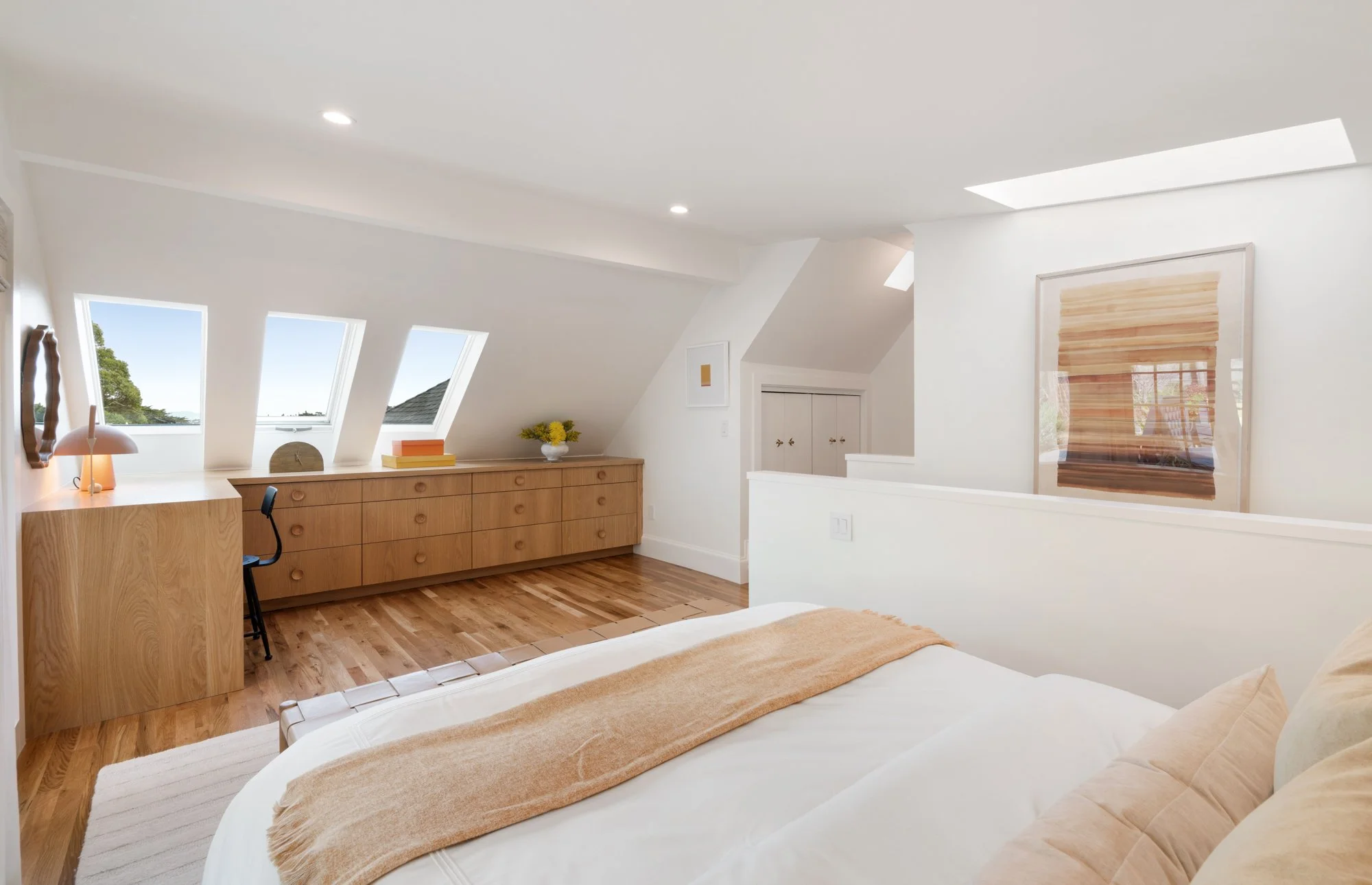 A bright bedroom with white walls, wooden flooring, a bed with beige bedding, a wooden dresser with three skylight windows above, and a small desk with a lamp and chair.
