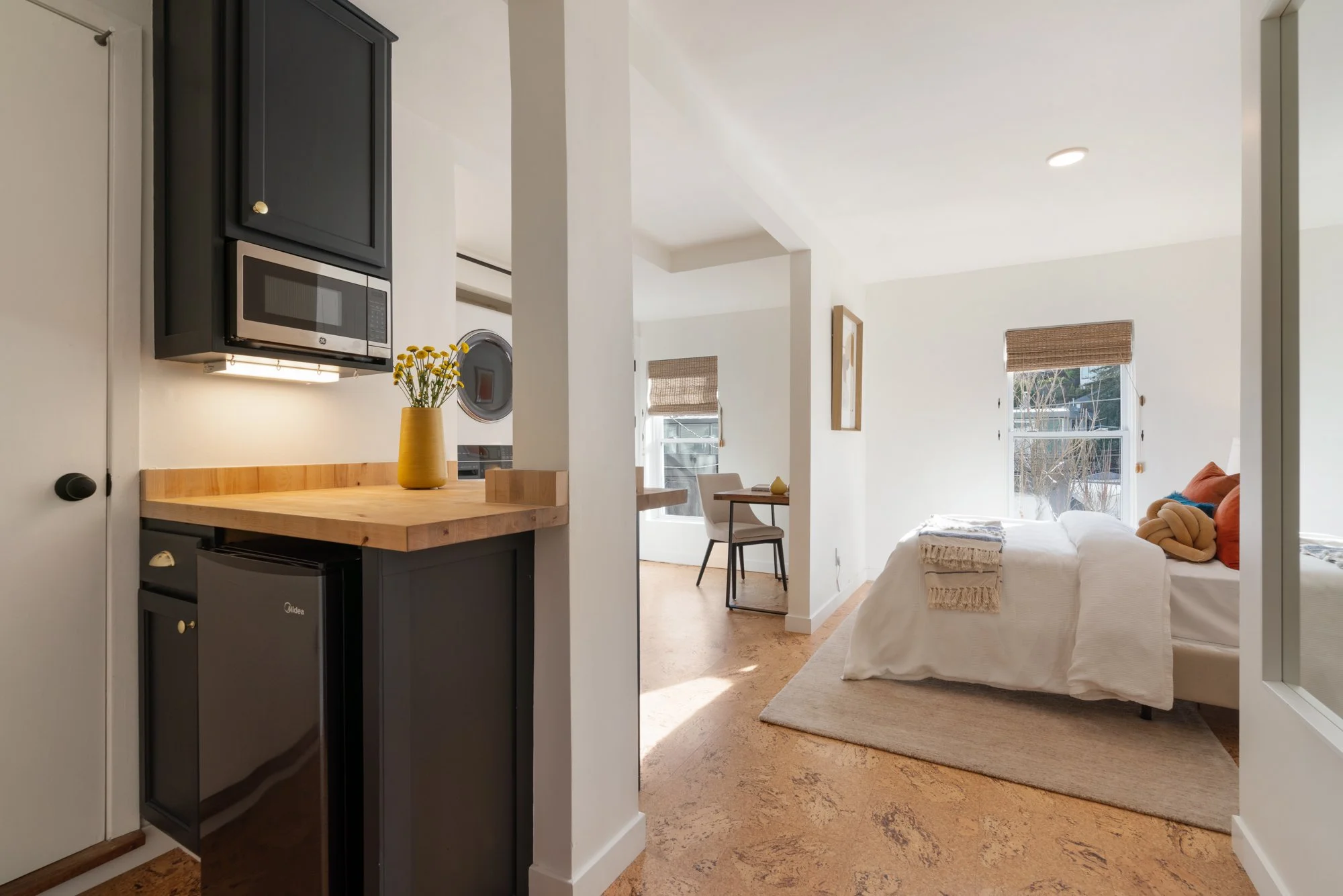 A cozy studio apartment with a kitchen nook, a dining area, and a bedroom with a bed, windows with woven blinds, and decorative pillows.