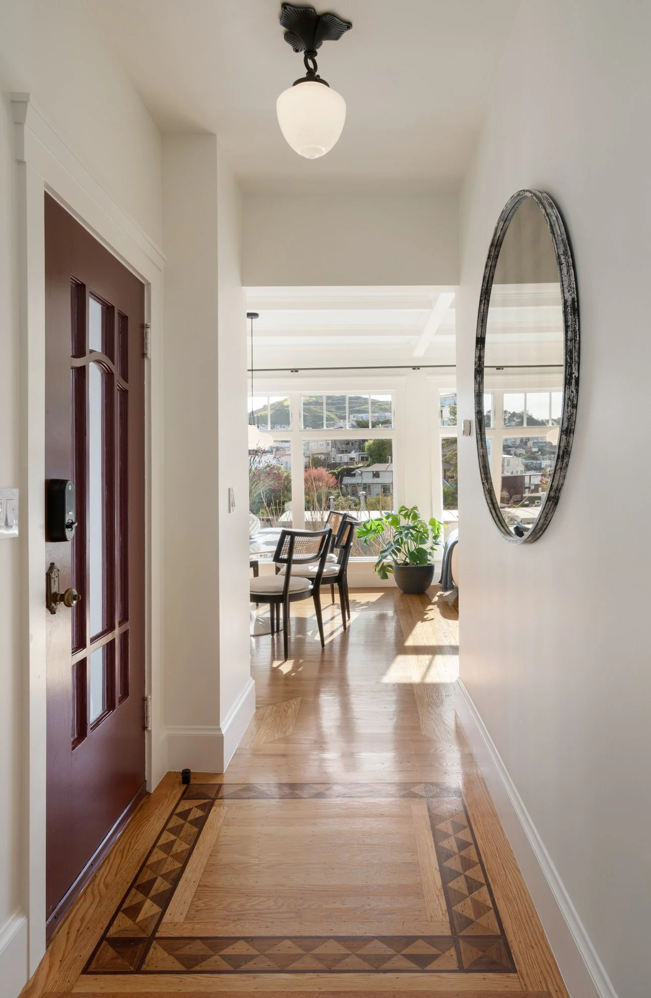 Hallway leading to a bright dining area, with a window view of houses and greenery outside, wooden flooring with decorative inlay, and a round mirror on the wall.