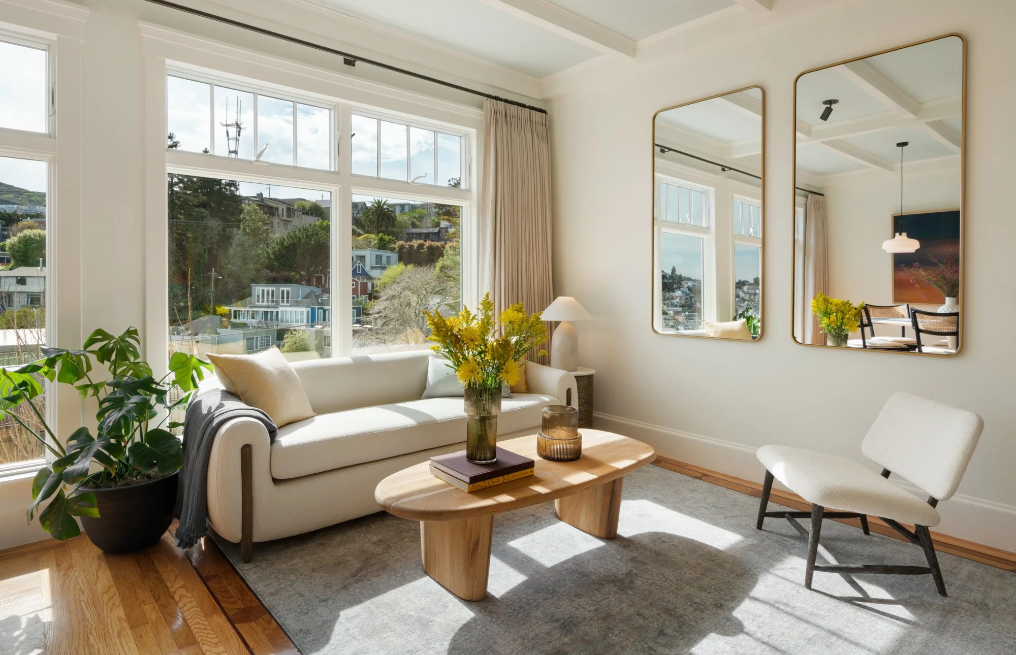 Bright living room with large windows, white sofa, wooden coffee table with yellow flowers, white armchair, and three rectangular mirrors on the wall.