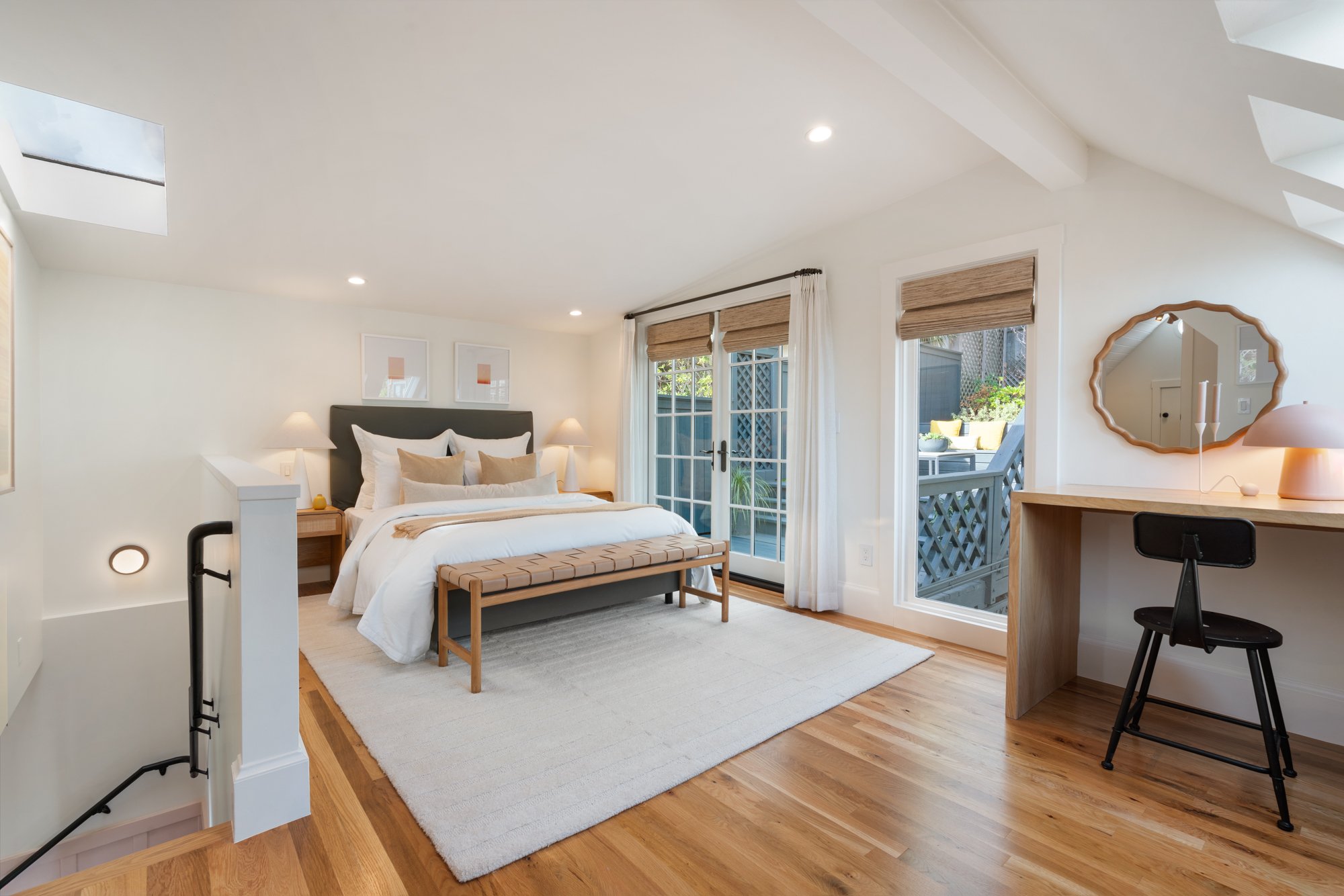 A bright bedroom with a large bed, nightstands with lamps, a sliding glass door and windows with brown blinds, wooden flooring, a bench at the foot of the bed, and a small workspace with a mirror and desk.