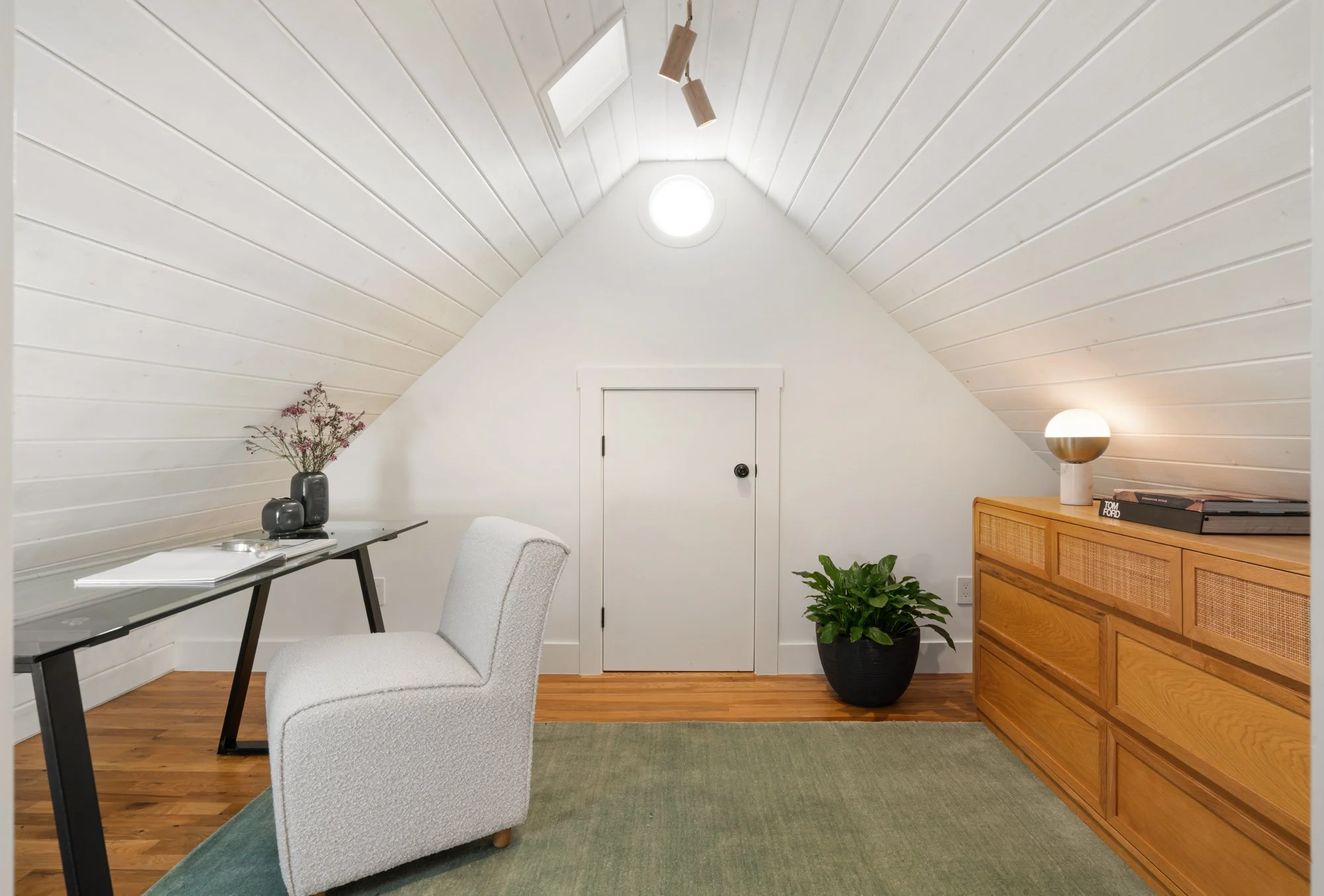 A bright, minimal attic room with sloped white wooden walls, a small door in the center, and wooden flooring. There is a simple glass desk with a white chair on the left, a potted green plant, a wooden sideboard with a lamp and some books on the righ