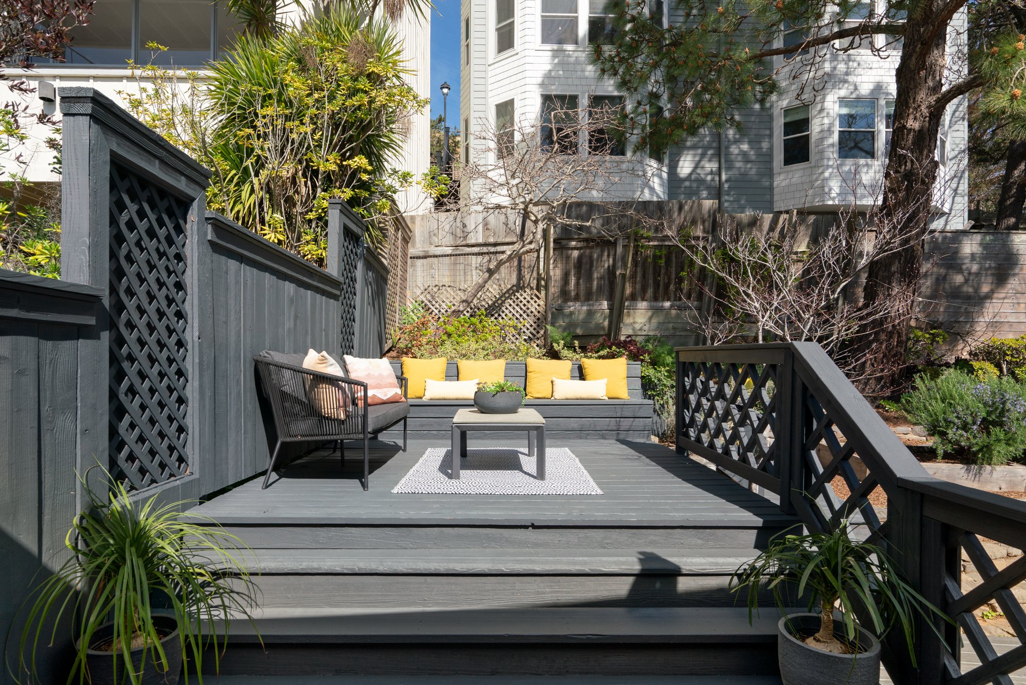 A cozy backyard patio with dark gray wooden decking, black wicker furniture, and decorative pillows. Potted plants and shrubs add greenery, and a white rug lies under a small coffee table. A built-in bench with yellow and white pillows is at the back