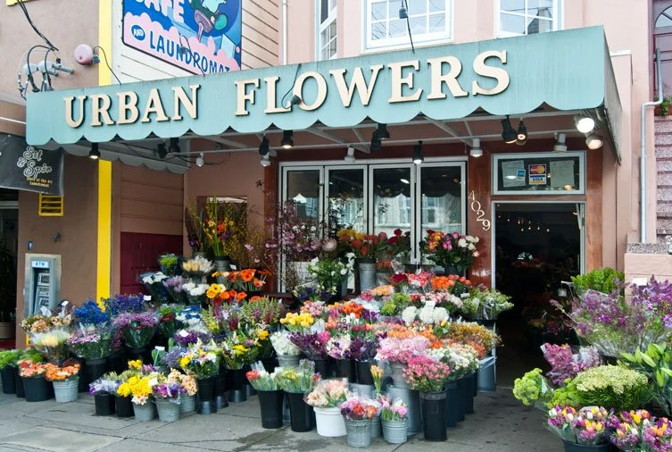 Street view of a flower shop named 'Urban Flowers' with colorful flowers displayed outside in buckets and pots.