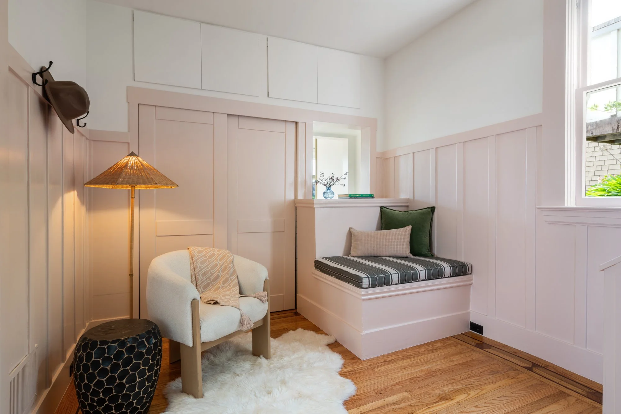Cozy room with white paneled walls, a small built-in window seat with striped cushion, green and beige pillows, a chair with a blanket, a standing lamp, and a side table with a honeycomb pattern design.