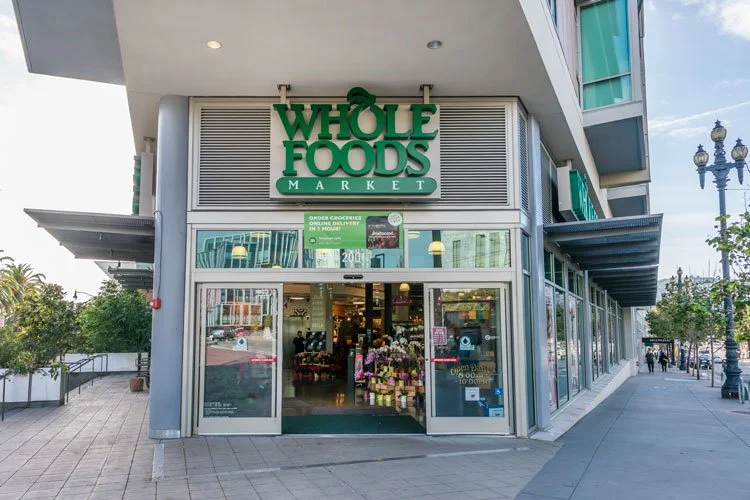 Exterior of Whole Foods Market grocery store on a city street, with large glass doors and window displaying flowers inside.
