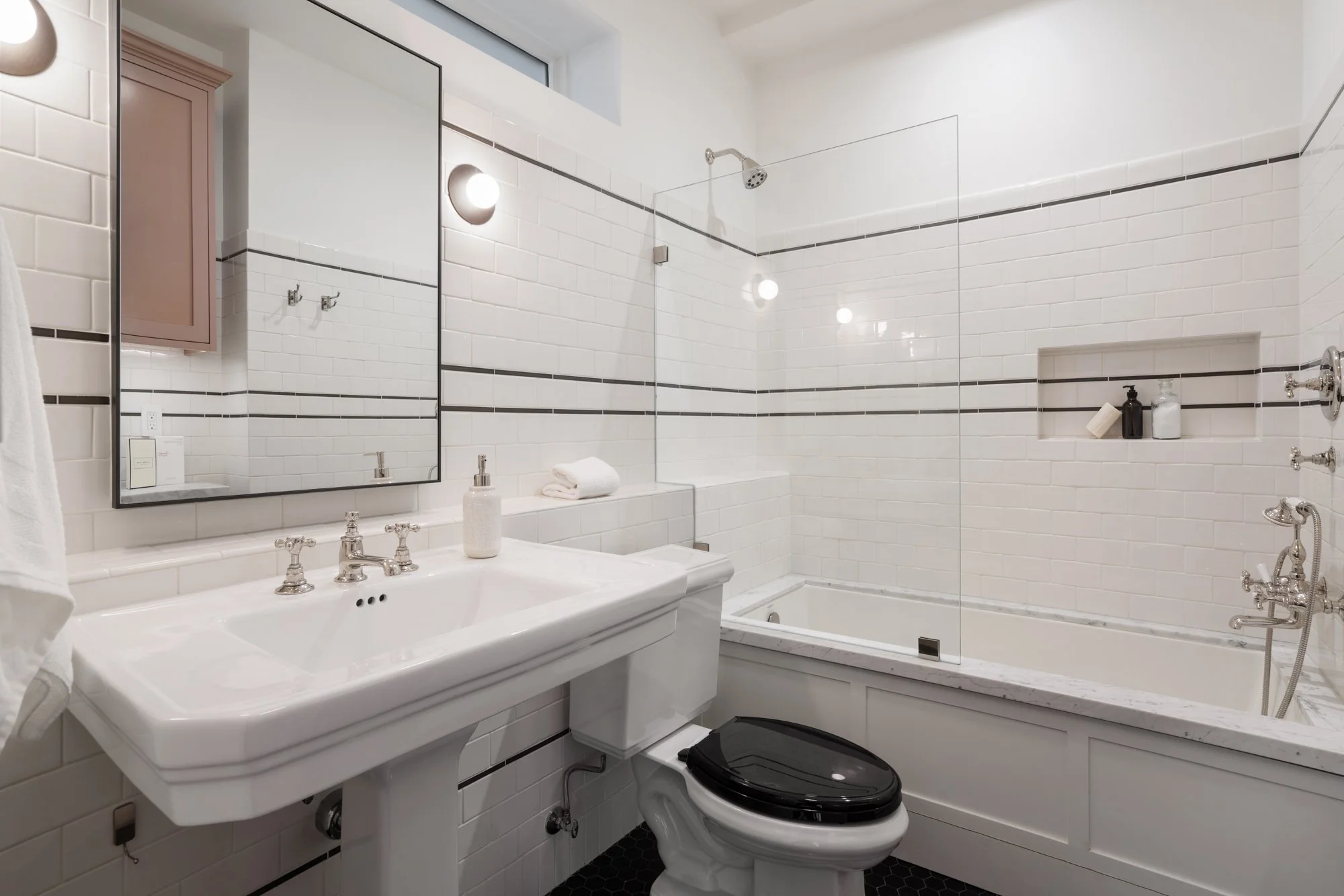 A clean bathroom with white subway tiles, a white sink with vintage-style fixtures, a rectangular mirror, a toilet with a black lid, a bathtub with a glass shower partition, and built-in wall niches with toiletries.