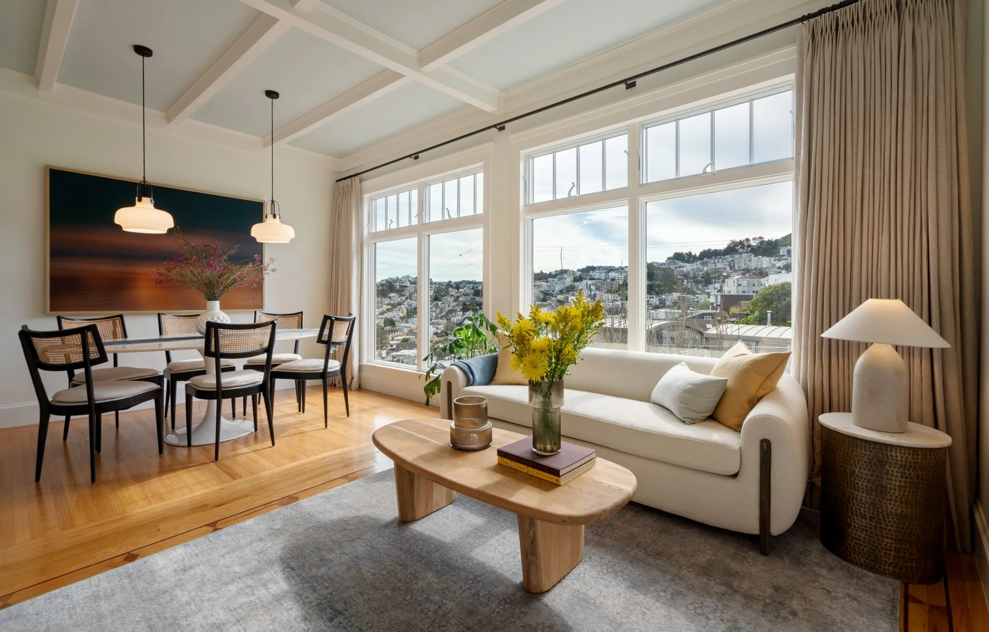 Living room with large windows, white sofa with pillows, wooden coffee table with a vase of yellow flowers, side table with lamp, dining area with a table and six chairs, wall art with sunset photo, and hardwood floors.