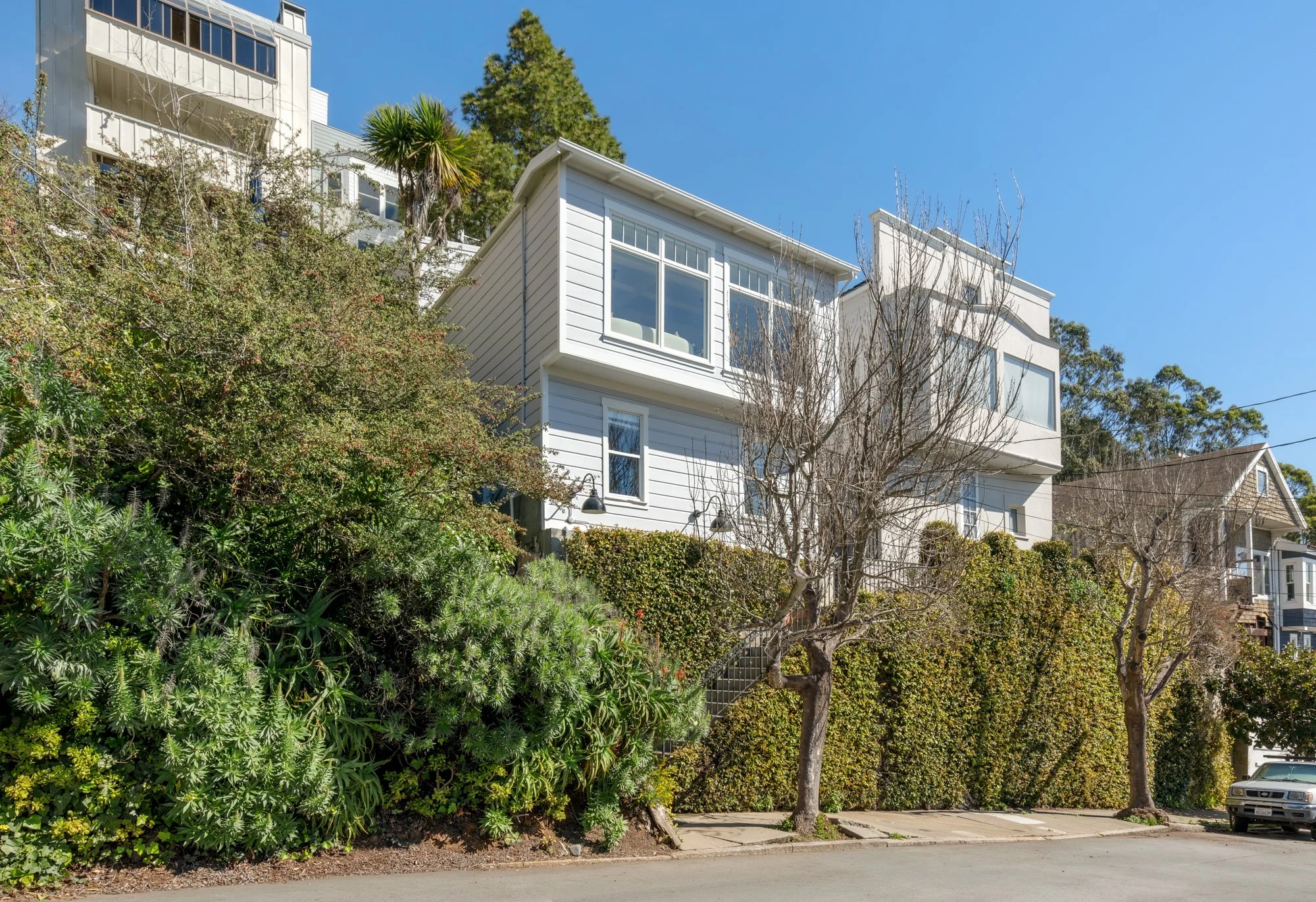 Multiple white residential buildings on a hillside with trees and shrubs in the foreground under a clear blue sky.