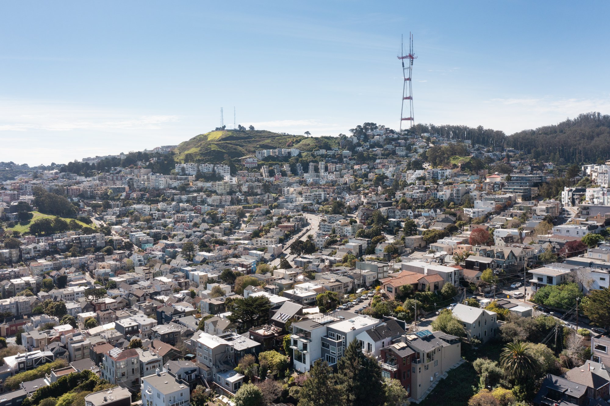 Aerial view of a city with numerous houses and buildings on hilly terrain, with a large radio tower on a hilltop in the background under a clear blue sky.