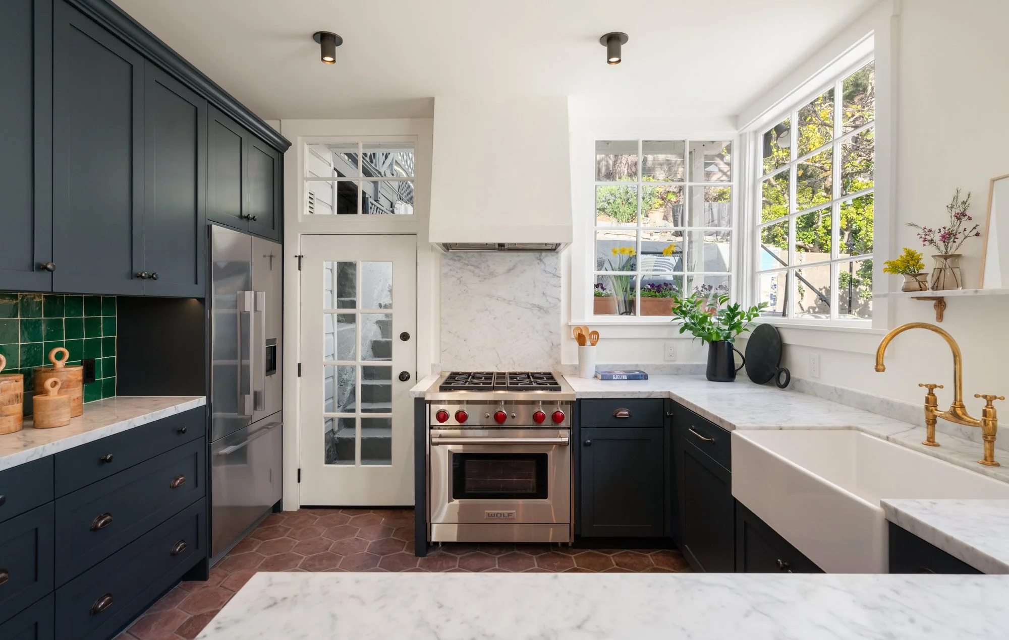 Modern kitchen with navy blue cabinets, white marble countertops, a white farmhouse sink with gold fixtures, large windows with a view of greenery, and stainless steel appliances, including an oven and refrigerator.