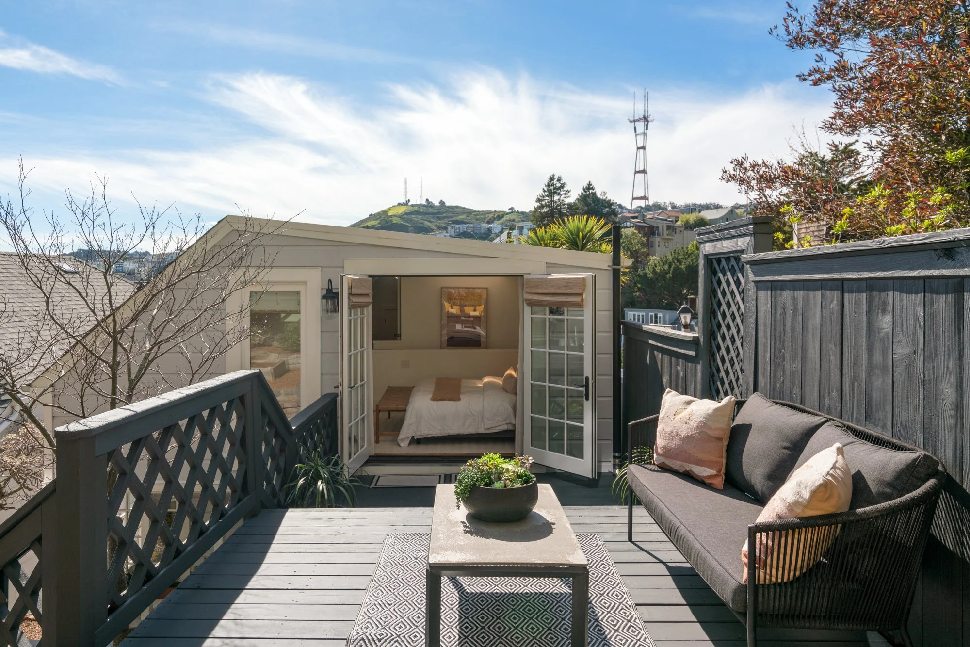 Outdoor balcony with black railing, gray wooden deck, black outdoor sofa with pillows, small rectangular table with a plant, and a tiny bungalow with open French doors showing a bedroom inside. In the background, green hills and a tall red and white 