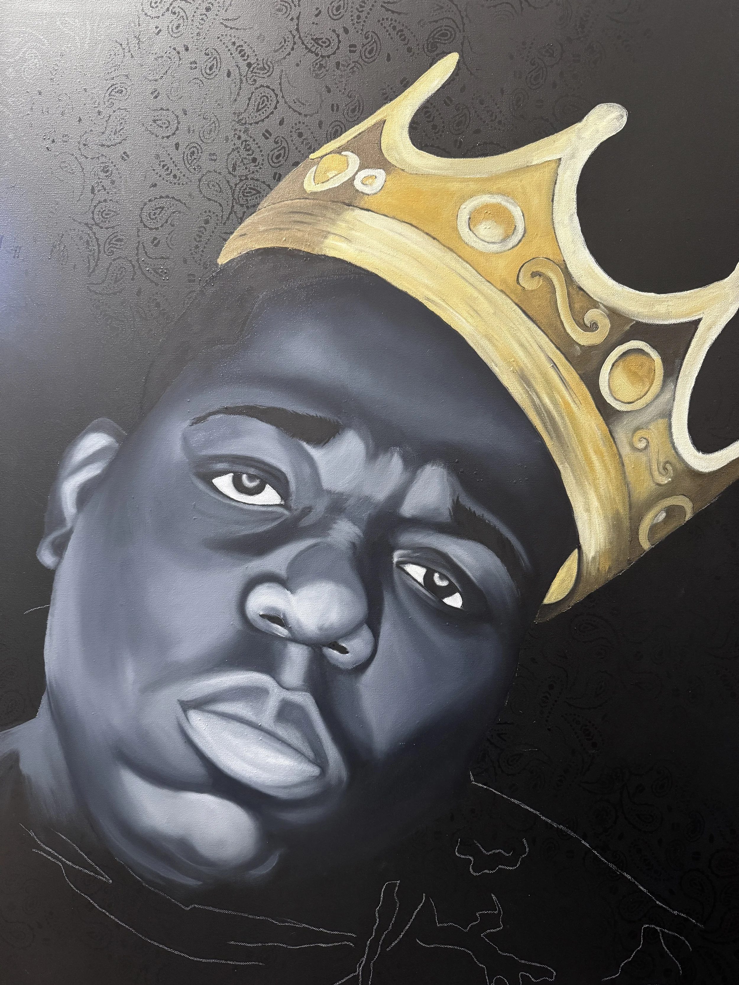 A black and white portrait of a young boy wearing a gold crown with decorative swirls, set against a dark patterned background.