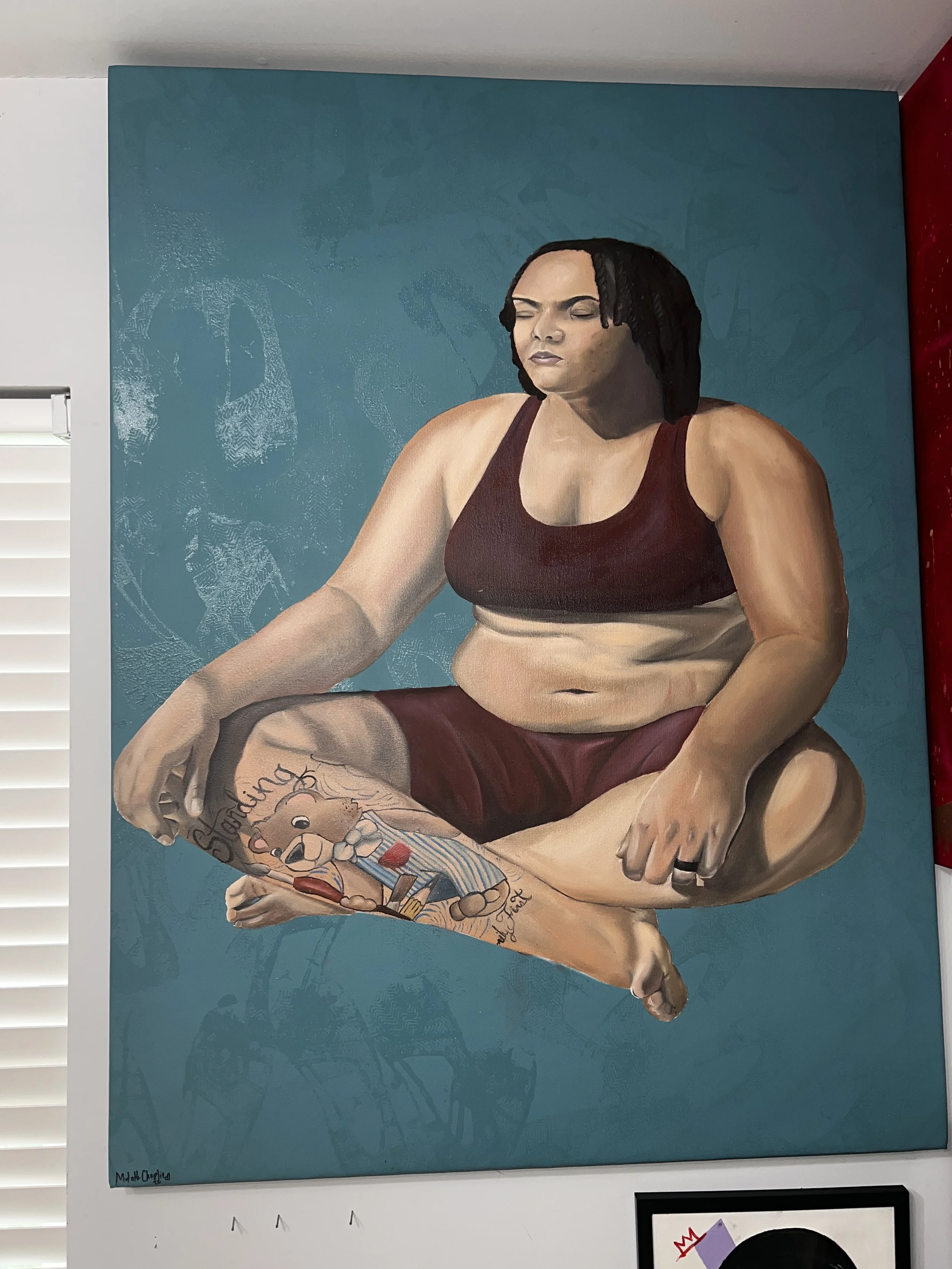 A mural of a woman sitting with legs crossed, wearing a dark sports bra and maroon shorts, with a tattoo of a cartoon character on her left leg, and a serious expression, set against a teal background.