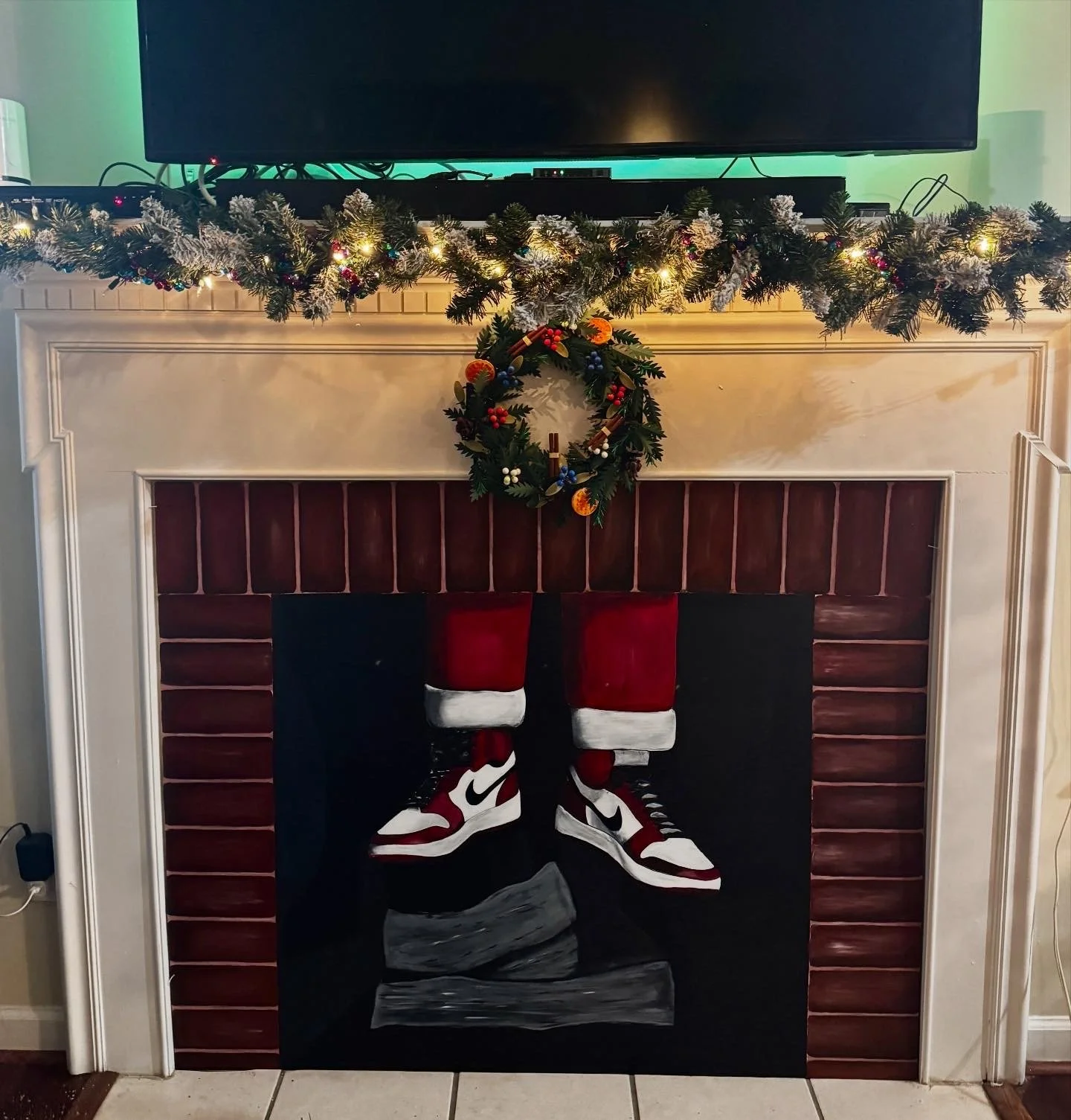 Painting of Santa's red and white sneakers hanging on a fireplace, decorated for Christmas with a wreath and garland.