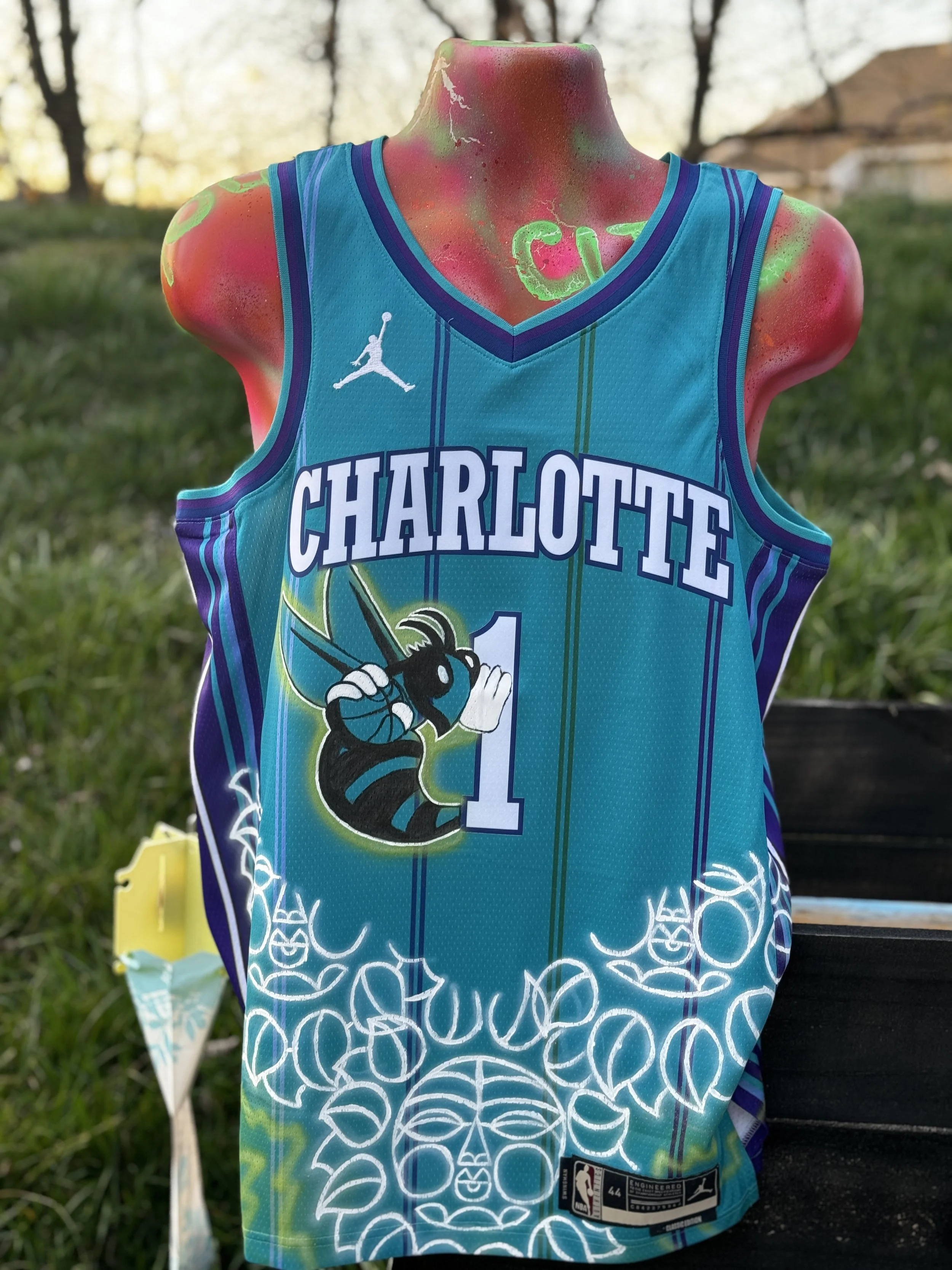 A mannequin wearing a Charlotte Hornets basketball jersey with the number 1, featuring the team's logo and colors, outdoors on grass with some trees and a sunset in the background.