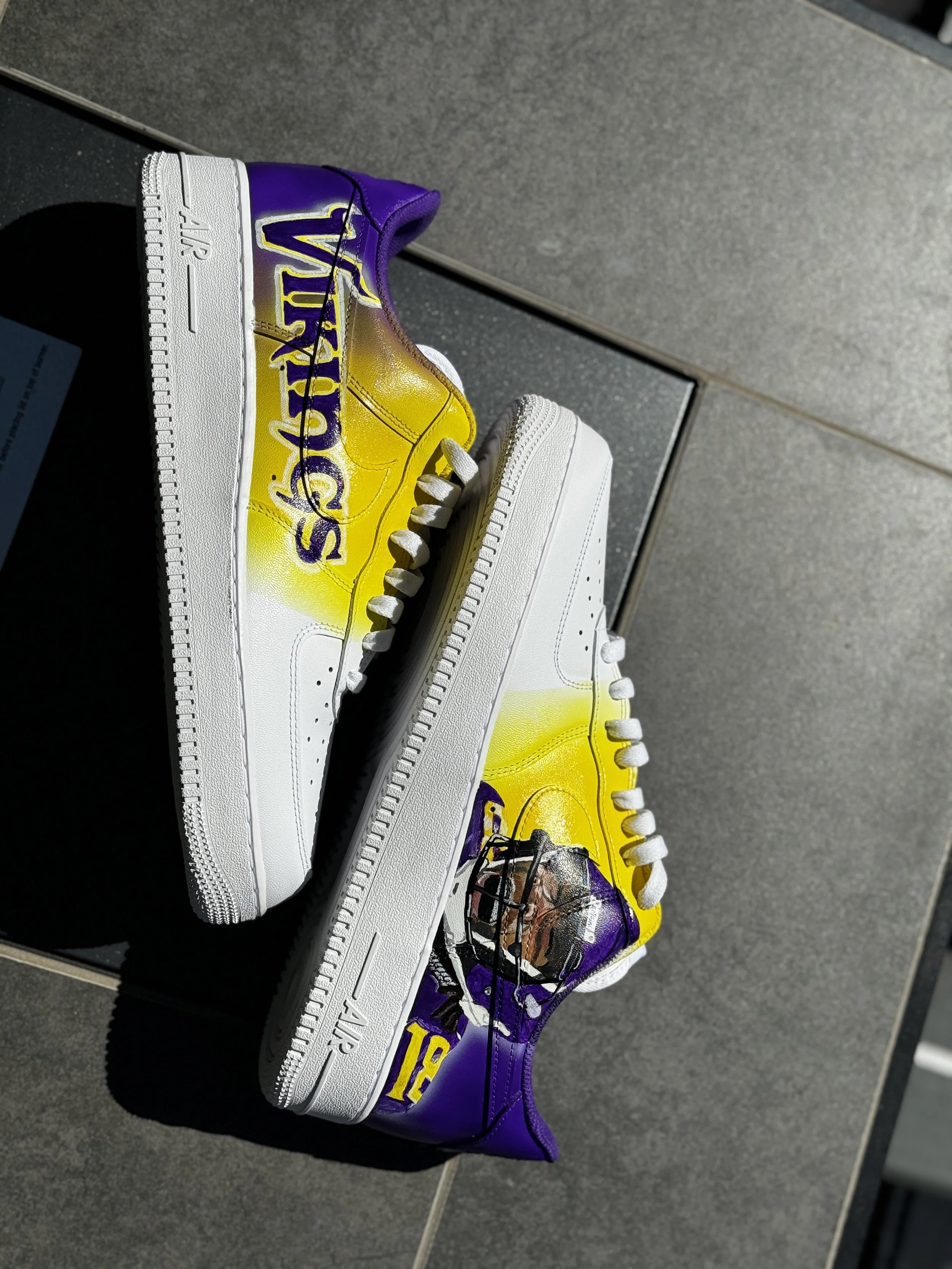 A pair of custom-designed Nike Air Force 1 sneakers with purple, yellow, and black paint, featuring the word 'Vikings' on the side and a football player illustration.