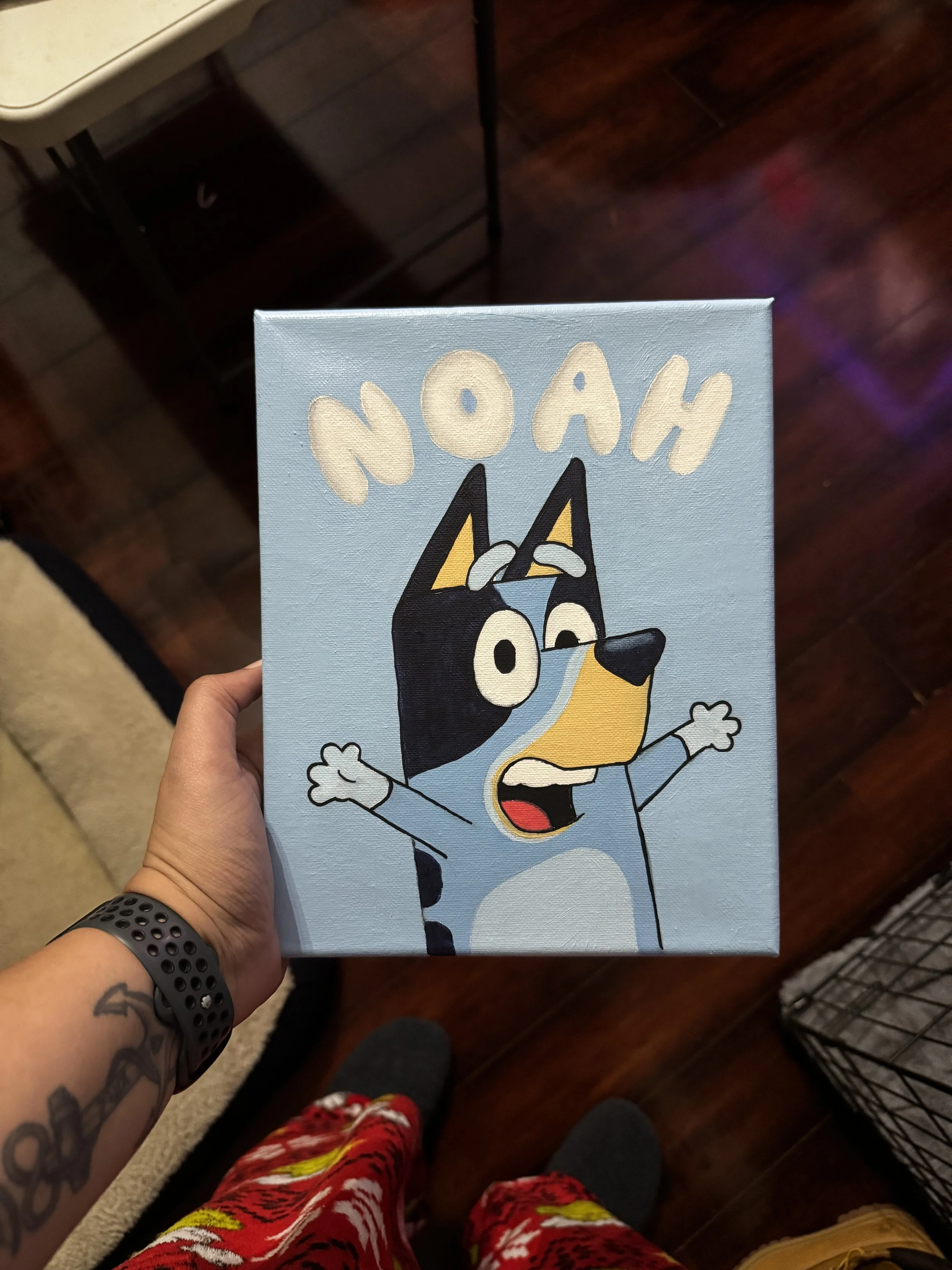 Hand holding a canvas painting of a cartoon dog with glasses, raising its arms, with the name "NOAH" written above.