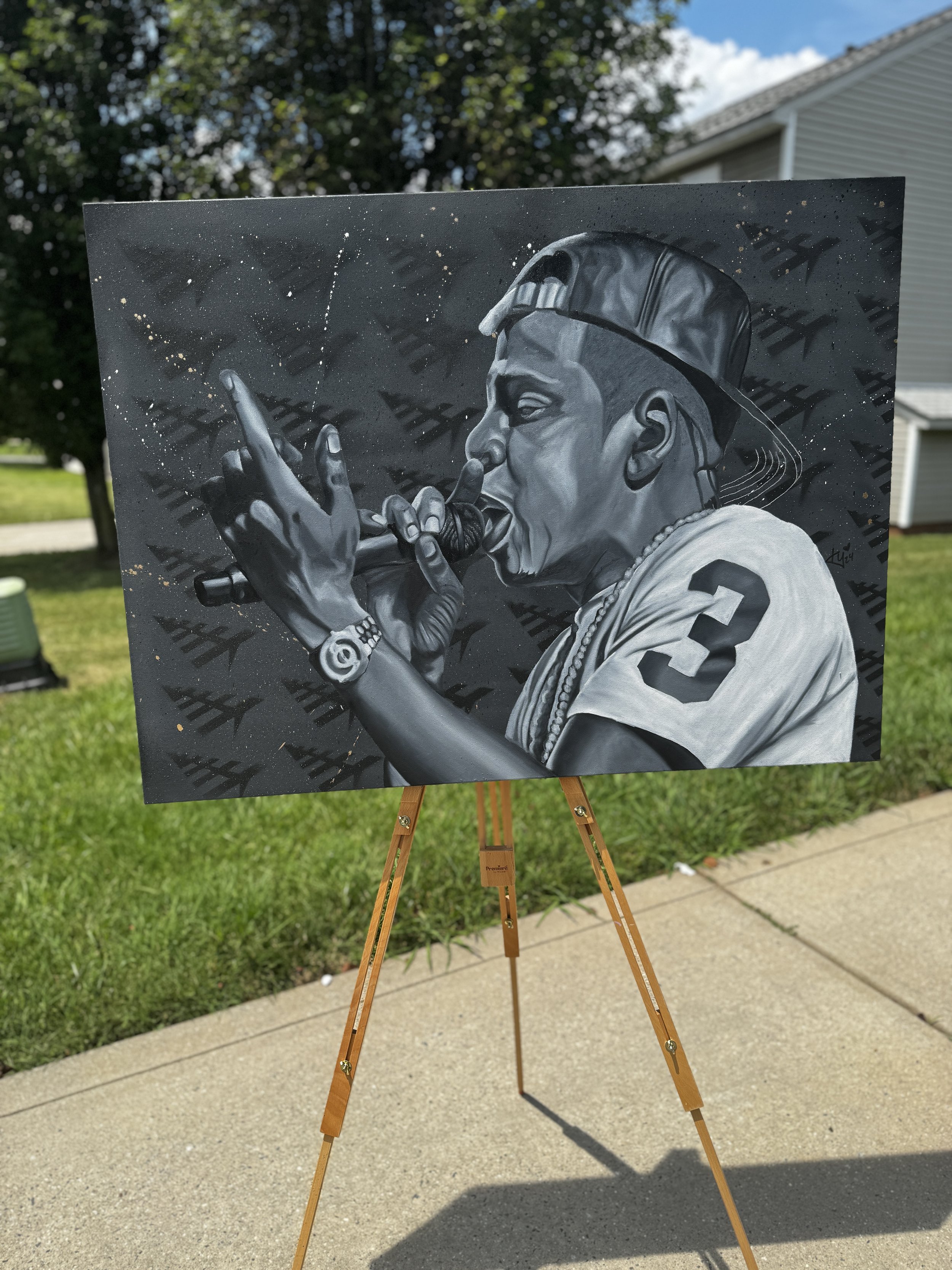 Black and white painting of a rapper holding a microphone, wearing a baseball cap backward, with a chain and wristwatch, displayed on a wooden easel outdoors.
