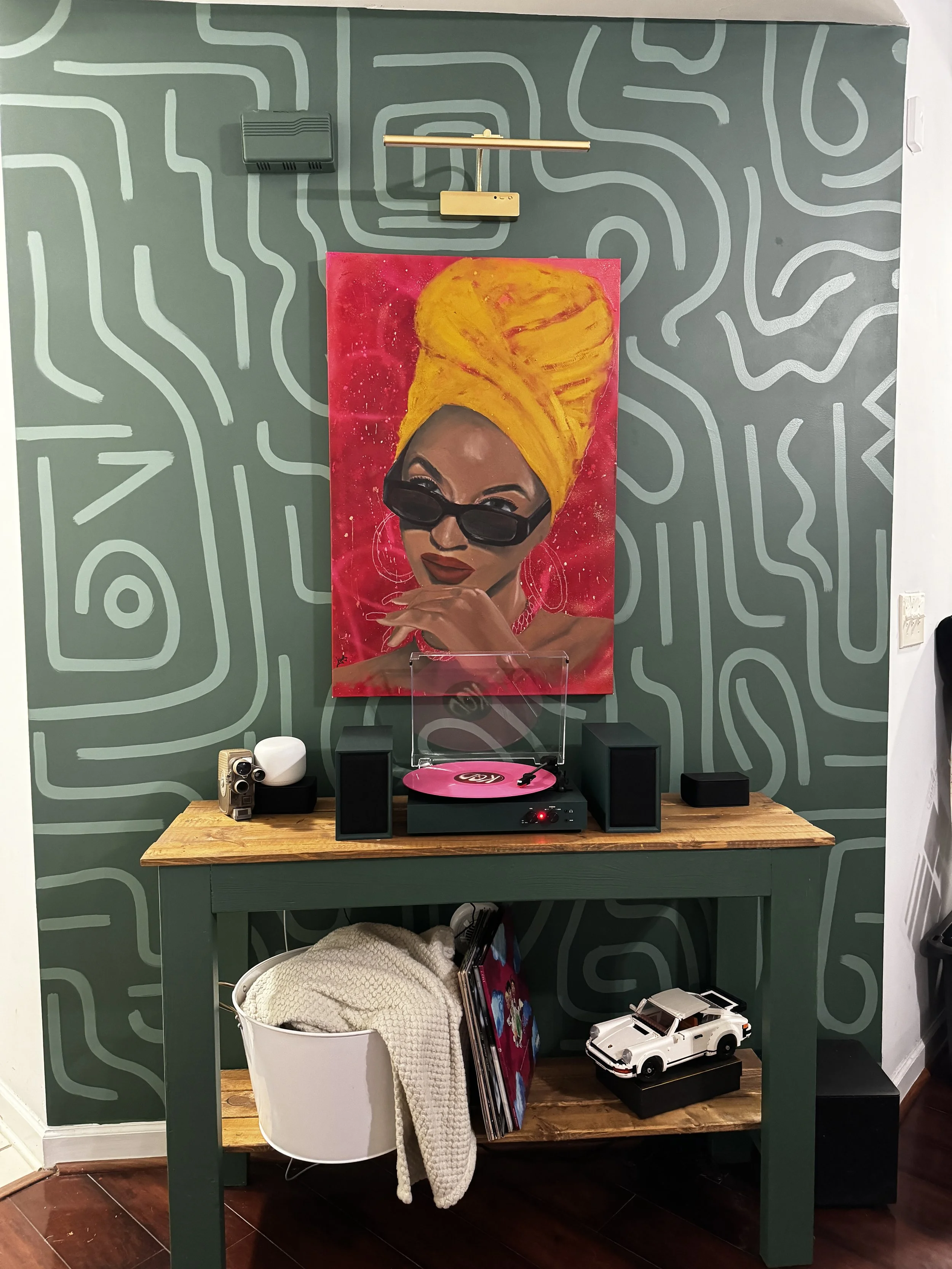 A music setup with a turntable on a green wooden table, a painting of a woman with a yellow headwrap and sunglasses hangs on the wall, and retro decor including a white toy car and vinyl records.