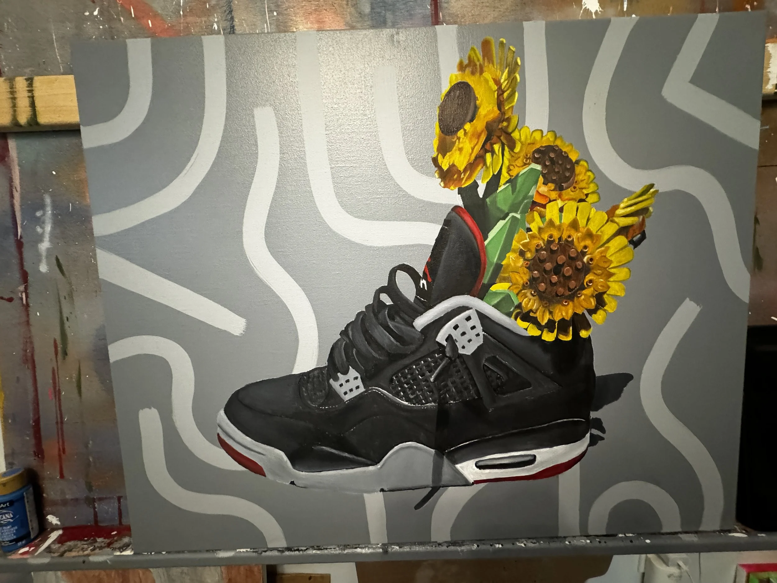 A painting of a black and white Air Jordan sneaker with three yellow sunflowers coming out of the shoe's opening, set against a gray background with white abstract lines.
