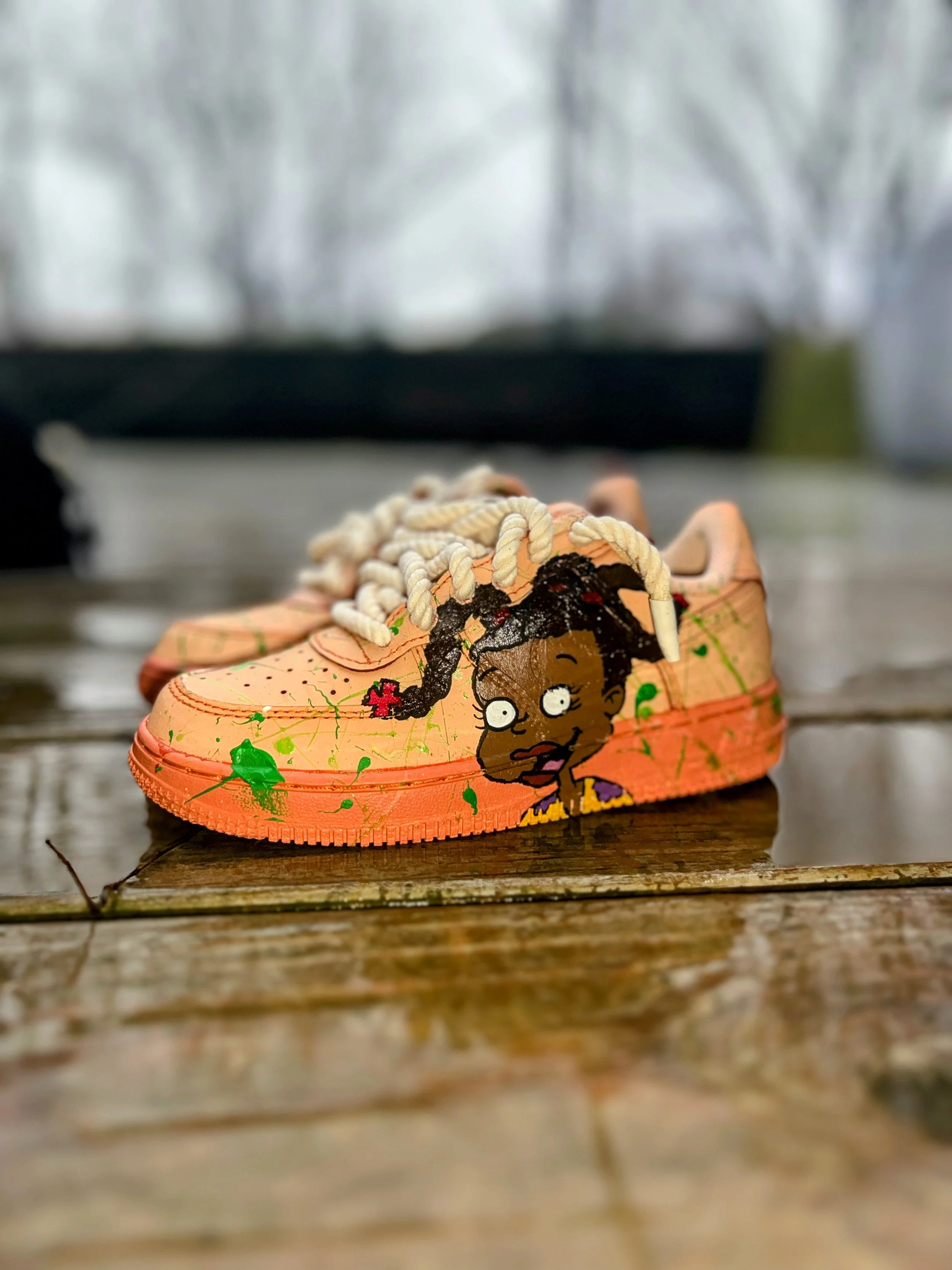 Pink sneaker with a cartoon girl painted on the side, featuring black hair, big eyes, and a surprised expression, with green and black splatters surrounding the character, placed on a wet wooden surface outdoors.