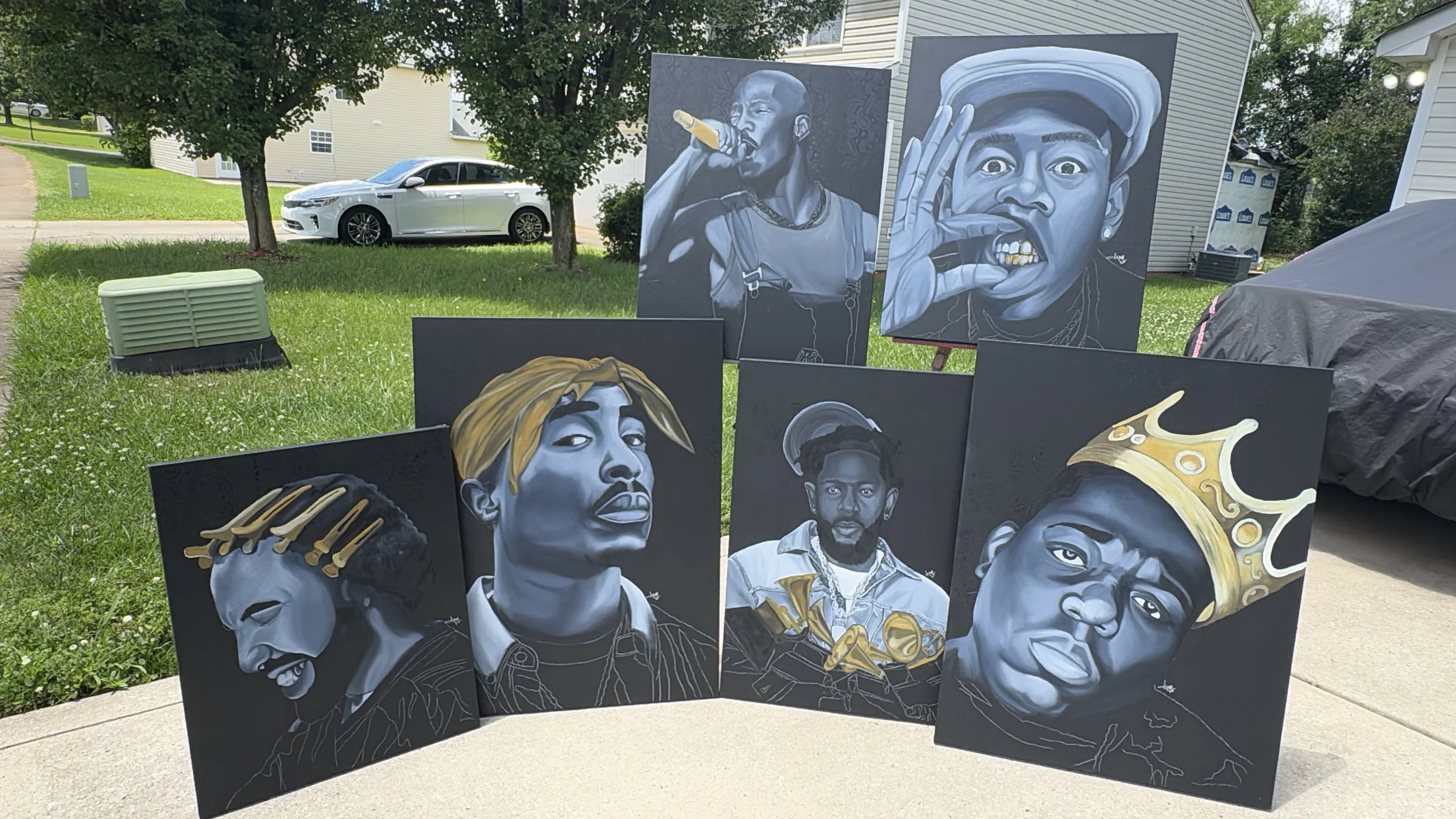 Six black and white portrait paintings of hip-hop artists displayed outdoors on a grassy area in front of houses, with a silver car in the background.