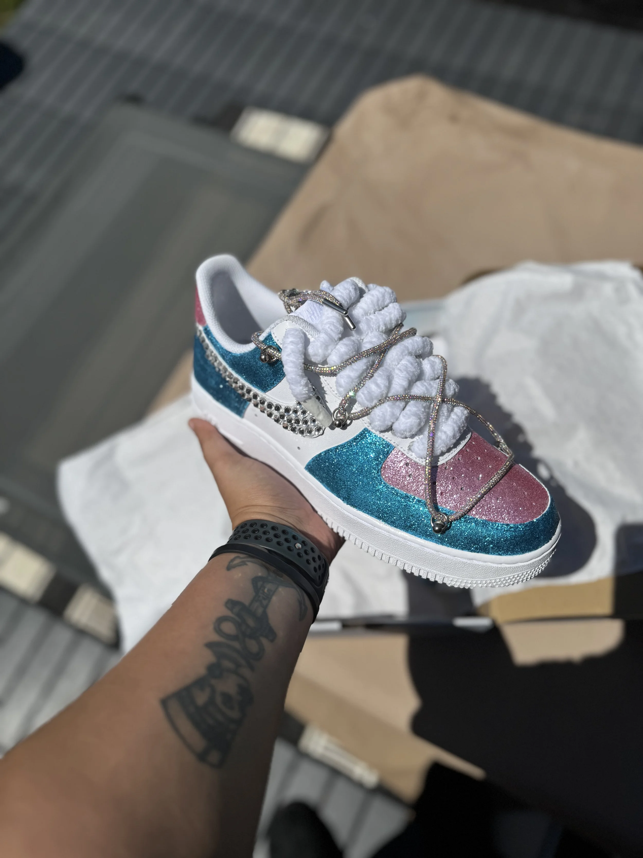 A colorful, glittery sneaker with pink, blue, and white sections, decorated with rhinestones and jewelry chains, being held by a person with a tattoo and a black wristband.