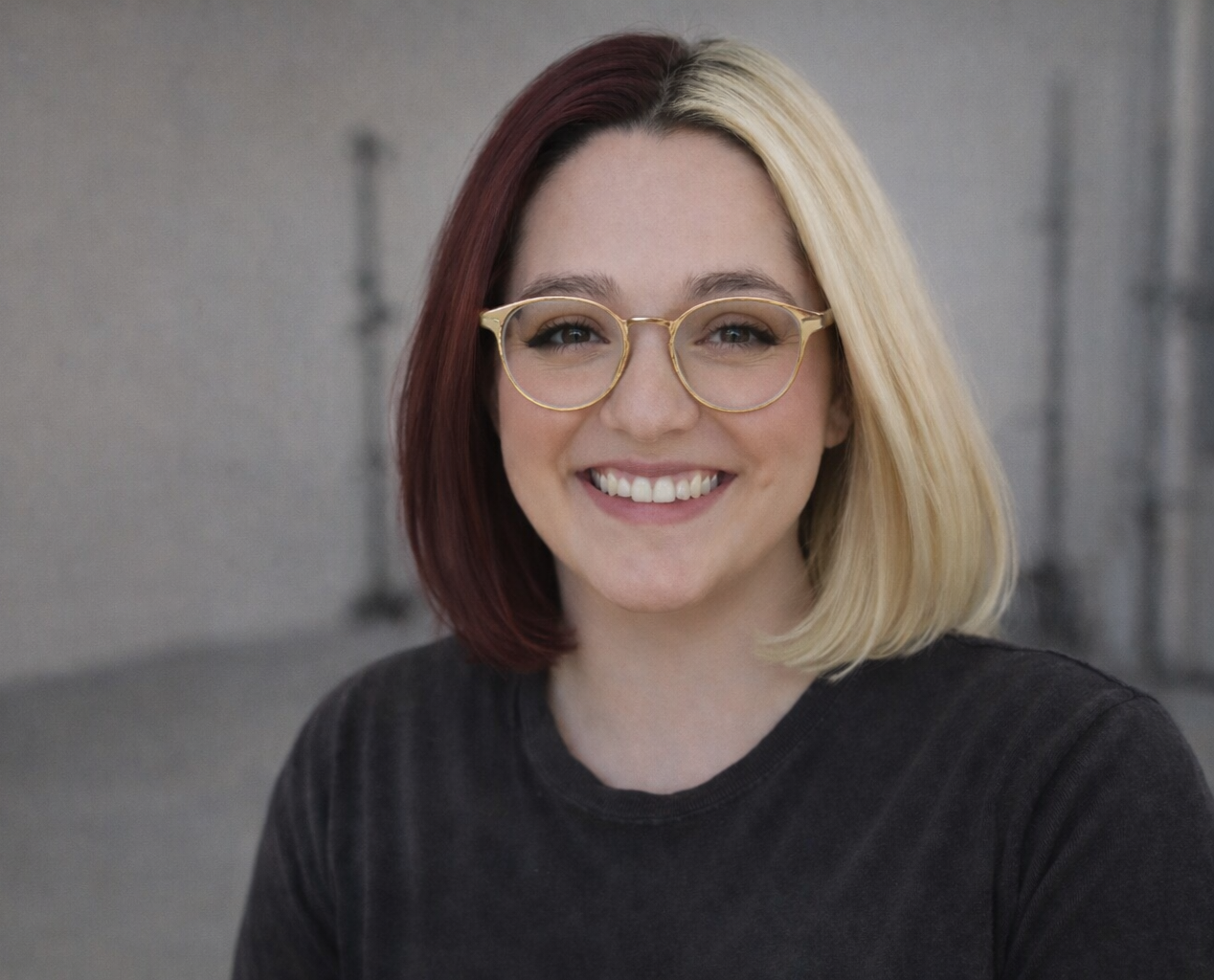 A young woman with shoulder-length hair, half dark red and half blonde, smiling and wearing clear-framed glasses and a black shirt, in front of a gray wall.