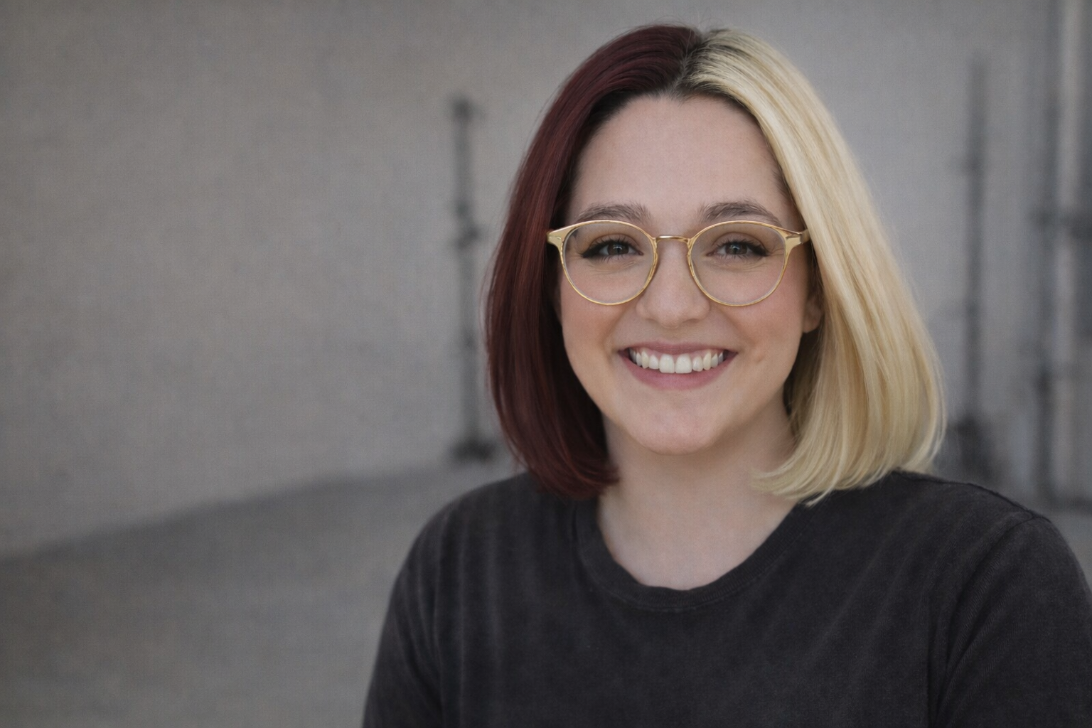 A young woman with shoulder-length hair, half blonde and half dark red, smiling and wearing round glasses and a black shirt.