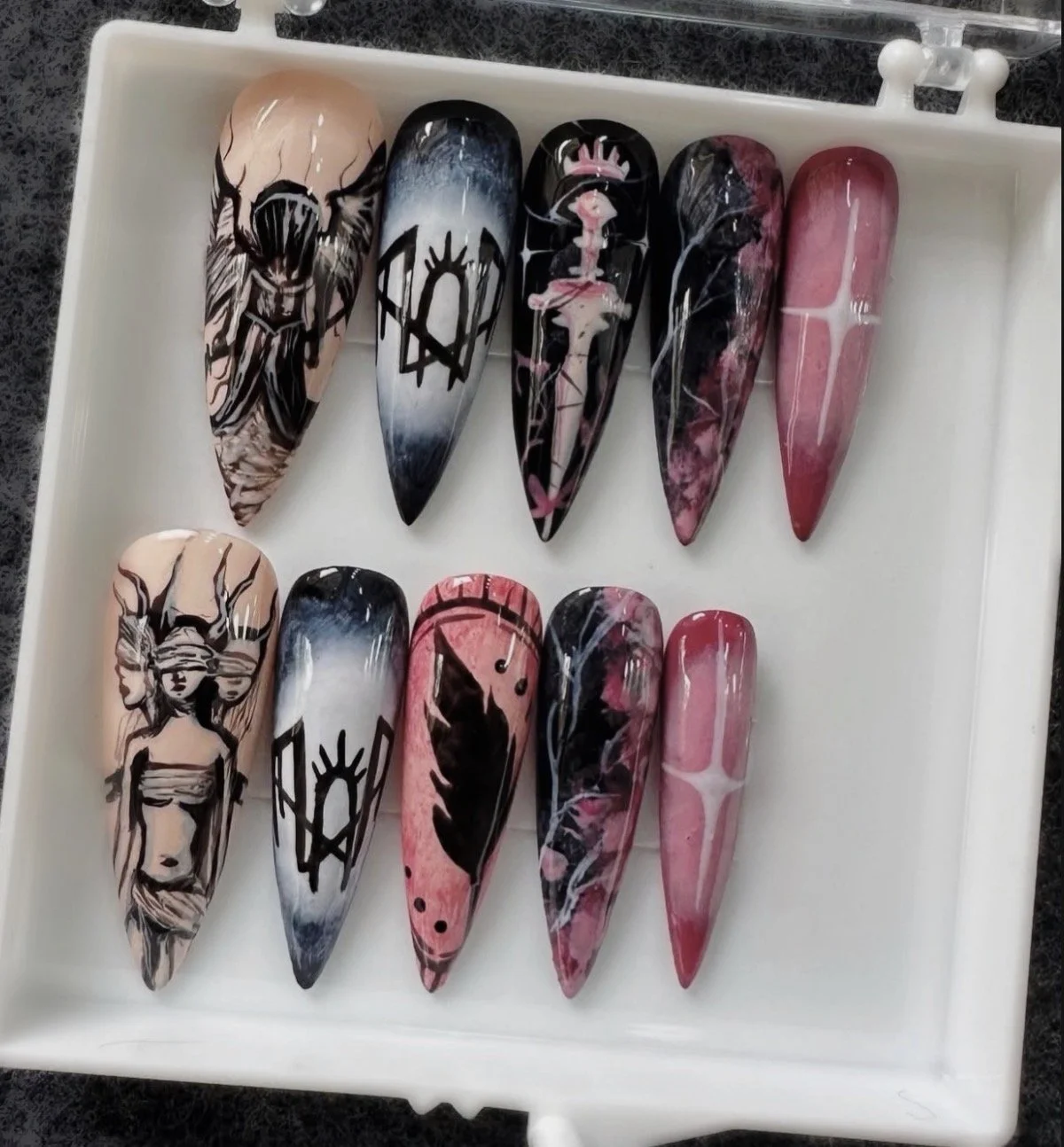 Display of ten decorated nail tips with artistic designs, including angels, abstract patterns, and illustrations in black, pink, white, and shades of blue.