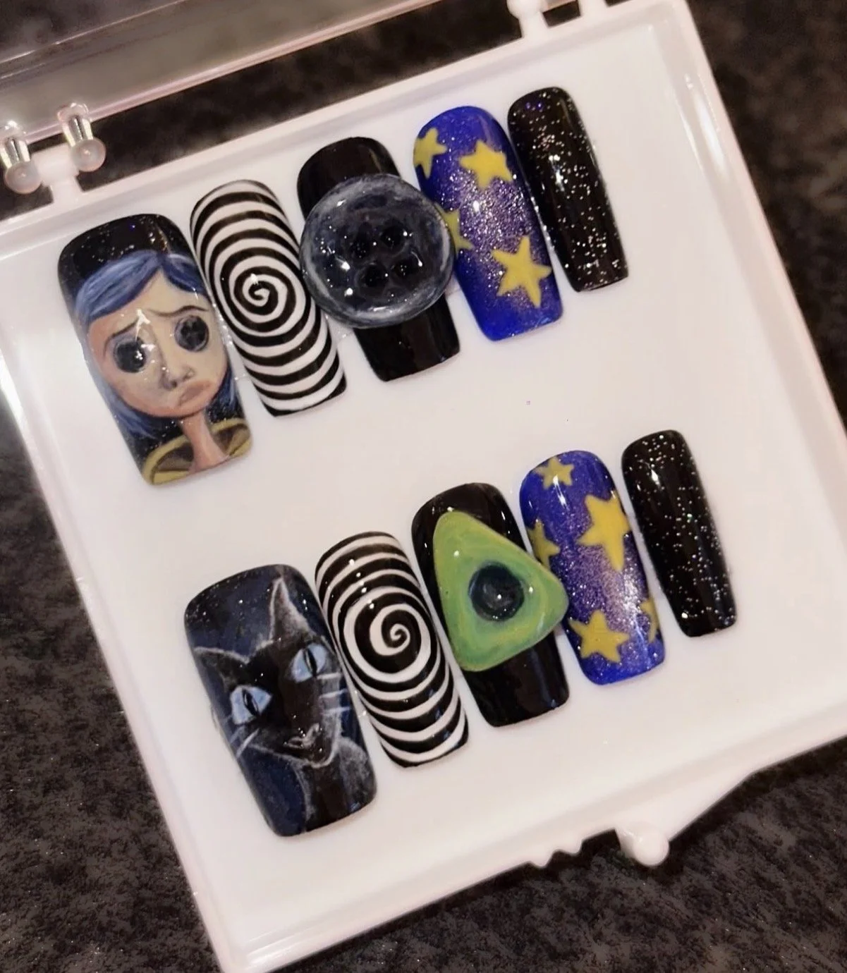 Set of decorative nail art designs with space-themed and abstract motifs, including a girl with blue hair, a cat face, spiral patterns, stars, and galaxy effects, displayed on plastic nail tips.