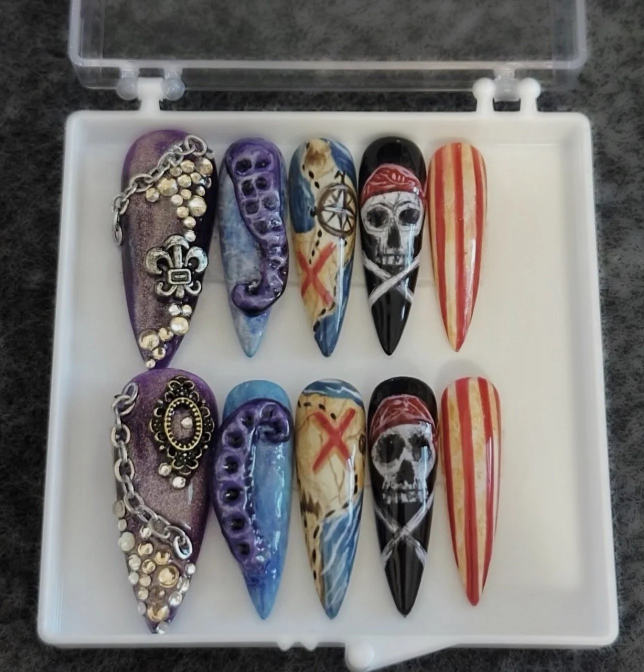 Two rows of decorated fingernails with pirate-themed and skull designs, some with chains, jewels, and painted stripes, in a white plastic case.